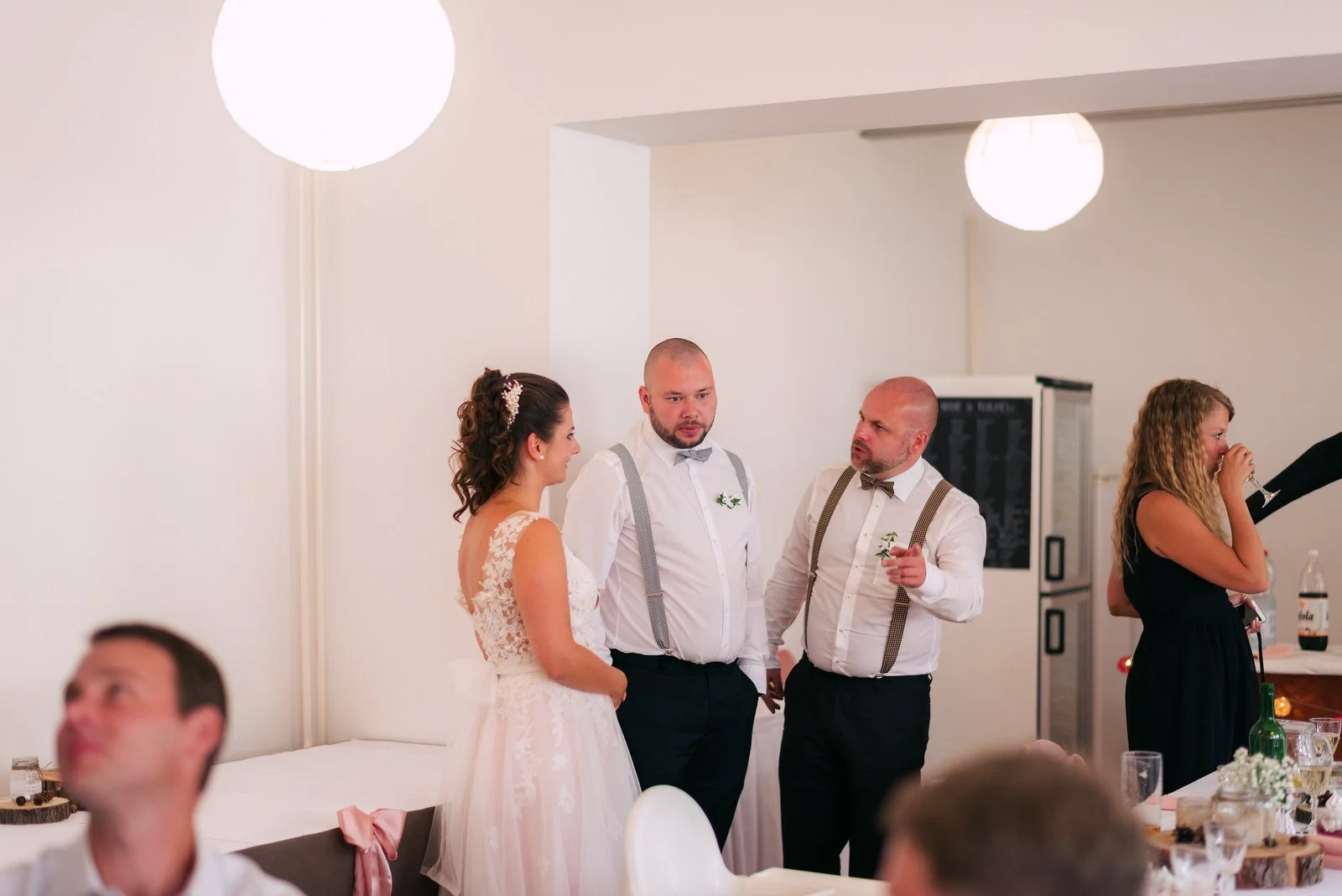 Žena stojí s mikrofonem před hosty a dvěma dalšími lidmi ve světlém sále.