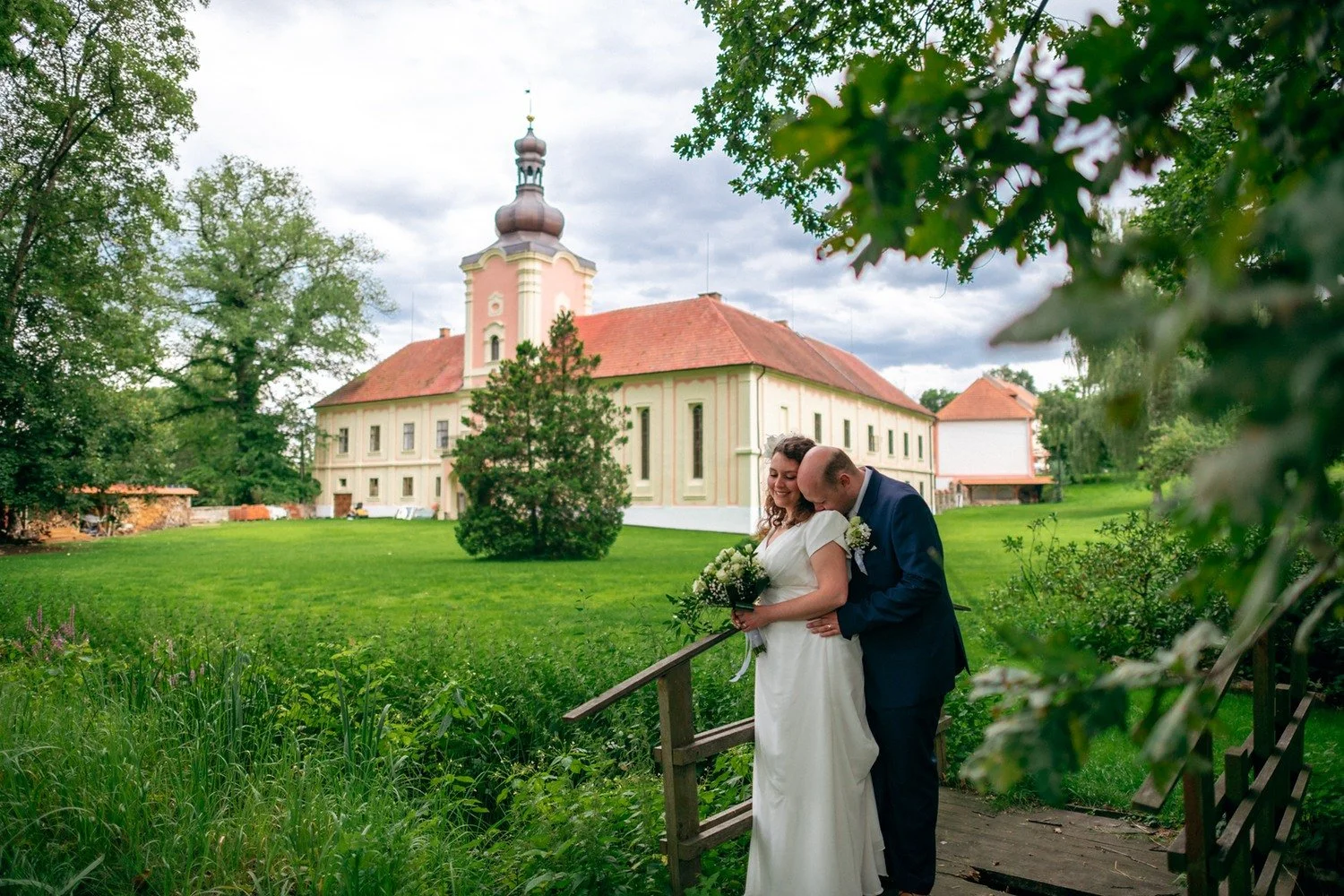 Nevěsta a ženich stojí v zeleni s areálem zámku v pozadí.