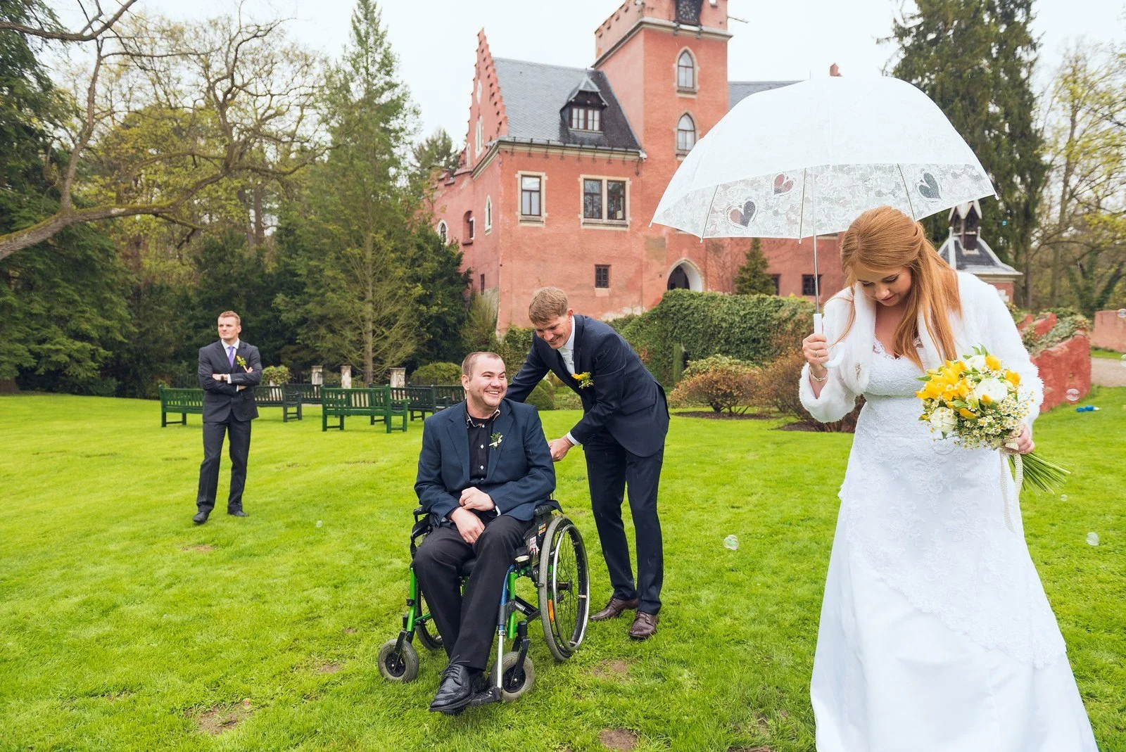Nevěsta s kyticí drží průhledný deštník, zatímco ženich klečí opodál na trávníku.