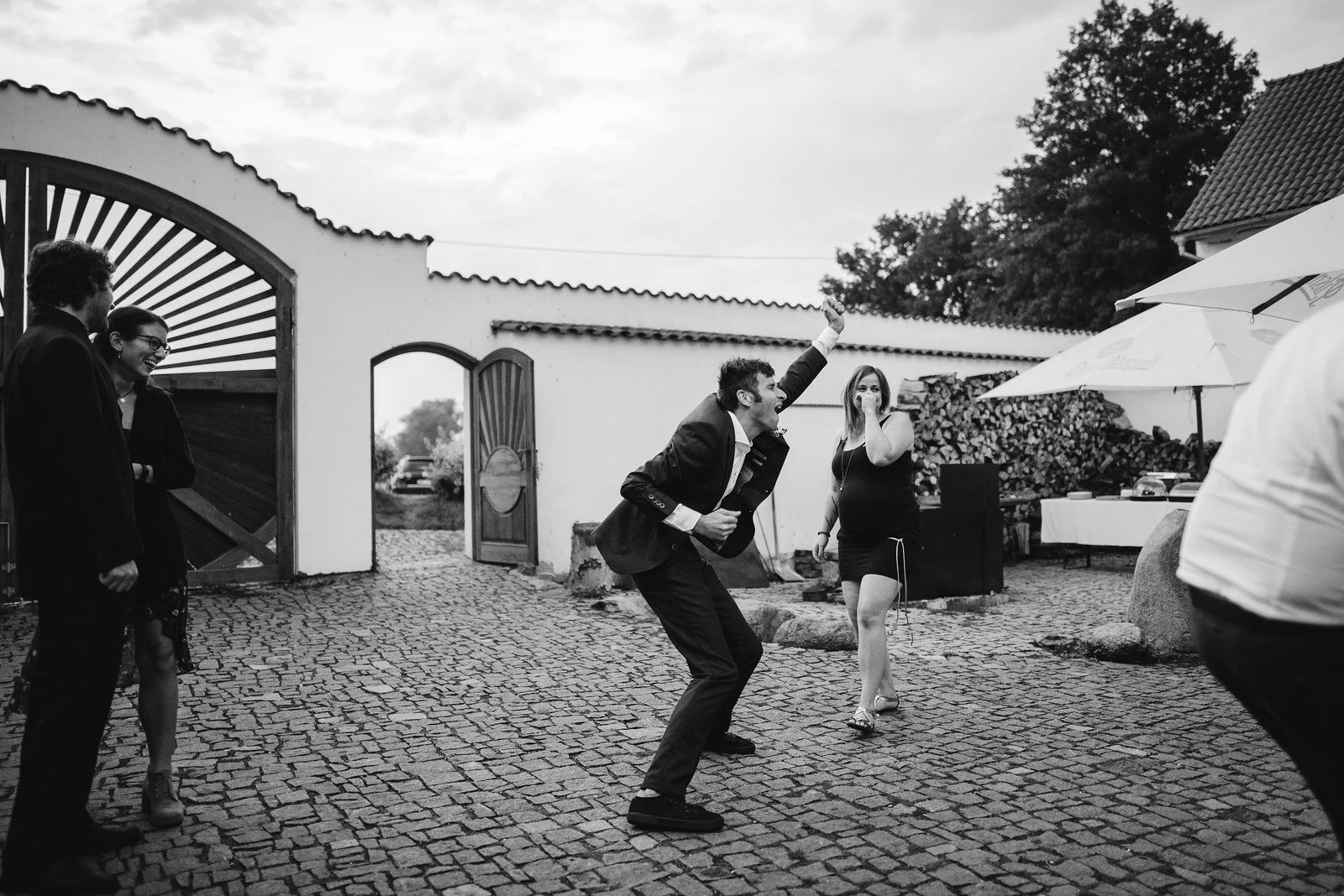 Černobílá fotografie ukazuje muže v podřepu při tanci na kamenném nádvoří.