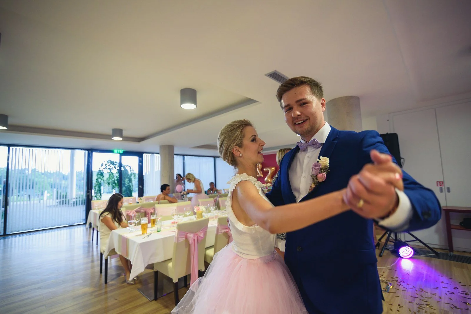Ženich tančí s ženou v růžové sukni uprostřed světlého sálu.