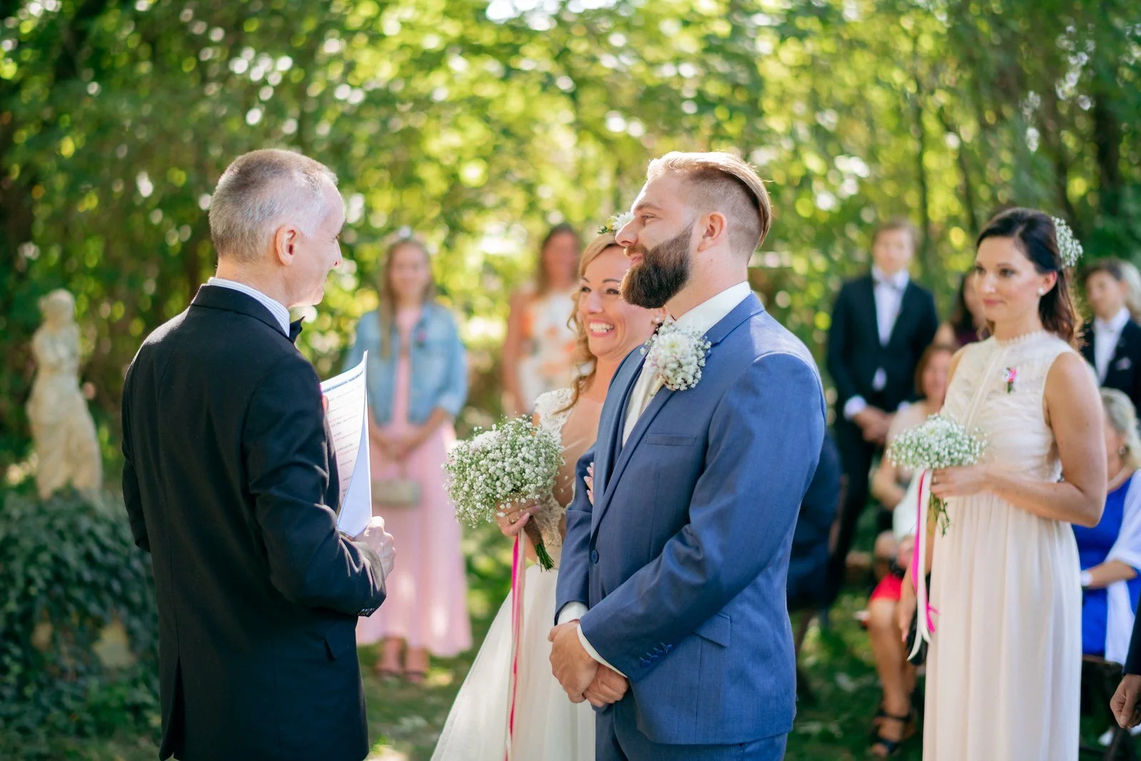Muž v modrém obleku drží ruku ženy během výměny prstenů před hosty.