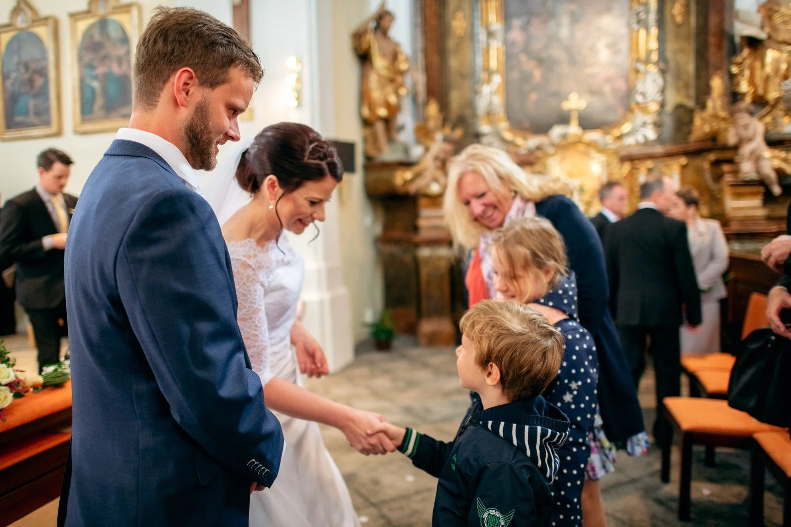 Muž v modrém obleku podává ruku chlapci v pruhovaném svetru, kolem něhož stojí další lidé.