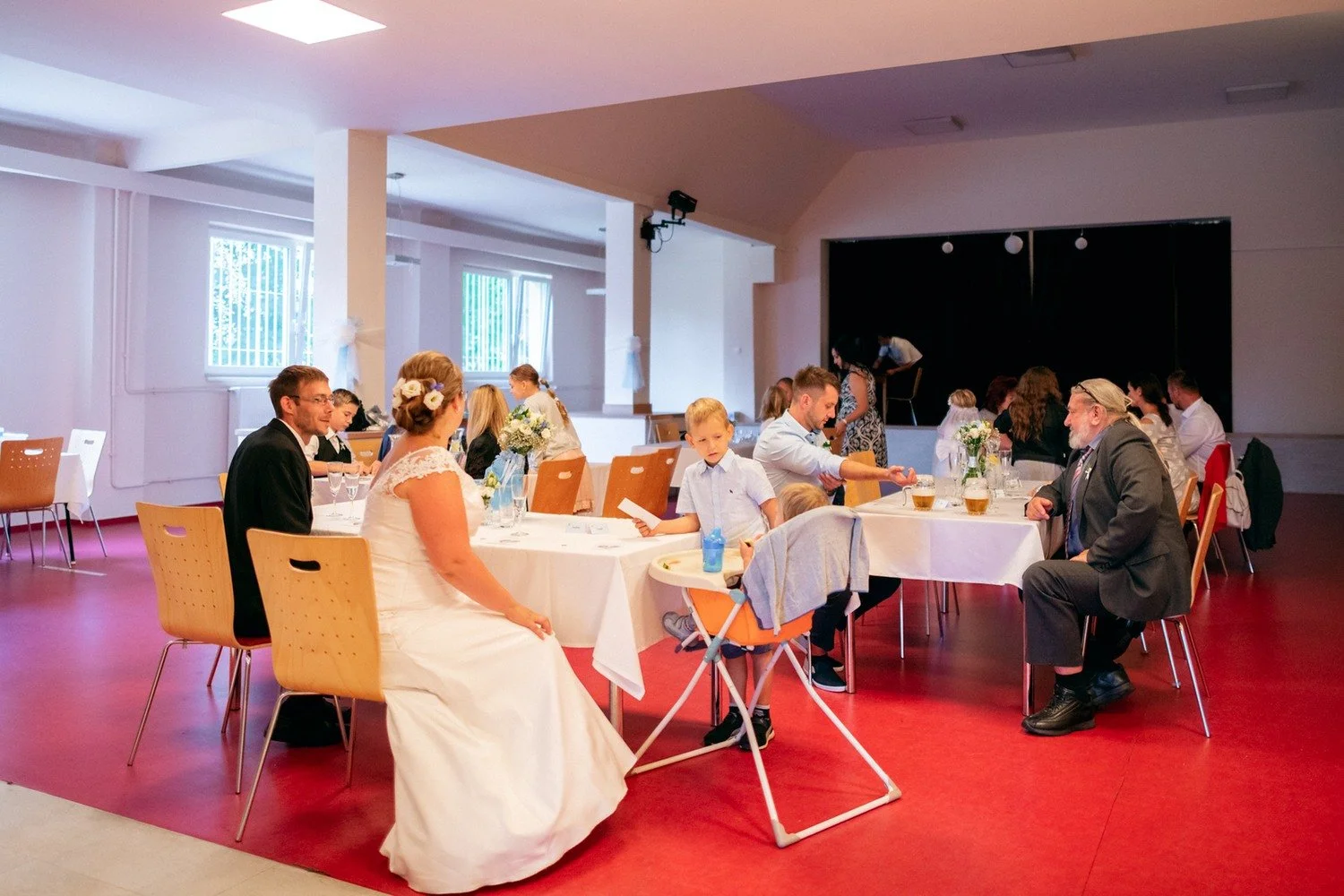 Hosté sedí u kulatých stolů v interiéru s červenou podlahou.
