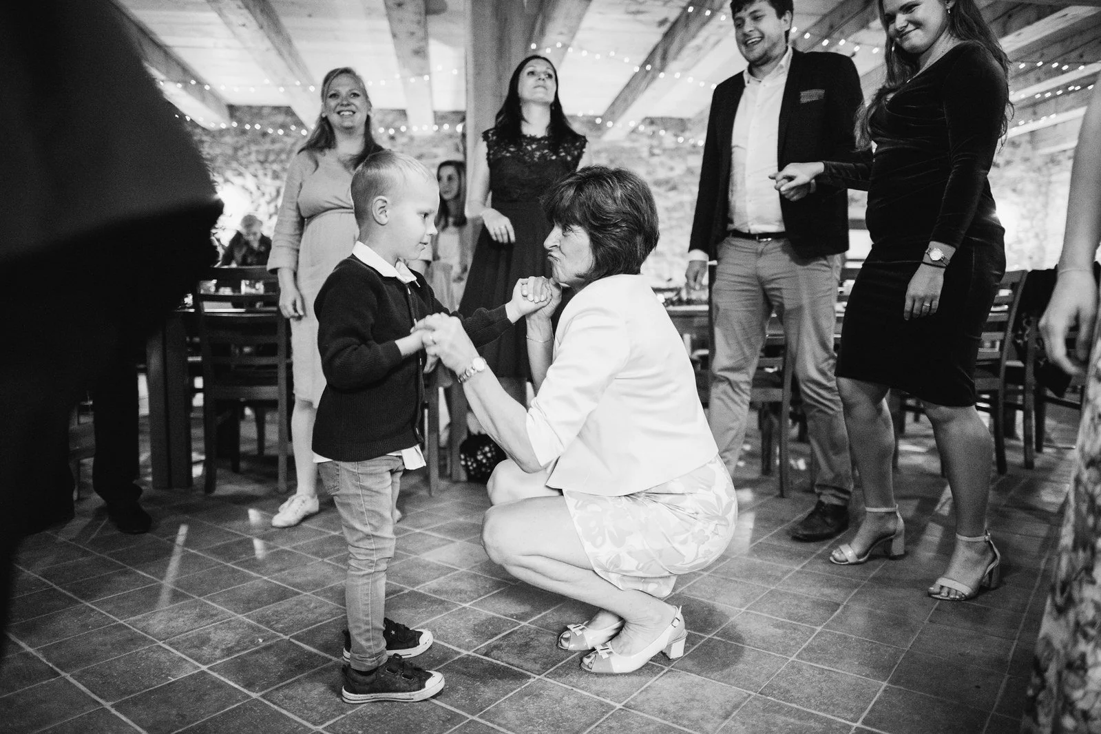 Černobílý snímek zachycuje klečící osobu ve světlém oděvu u malého chlapce mezi stojícími hosty.