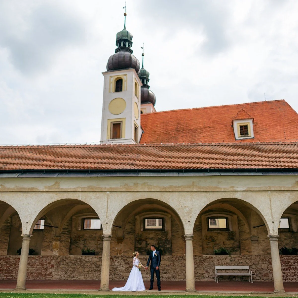 Jitka a Lukáš stojí pod podloubím, nad jehož červenou střechou vystupuje kostelní věž.