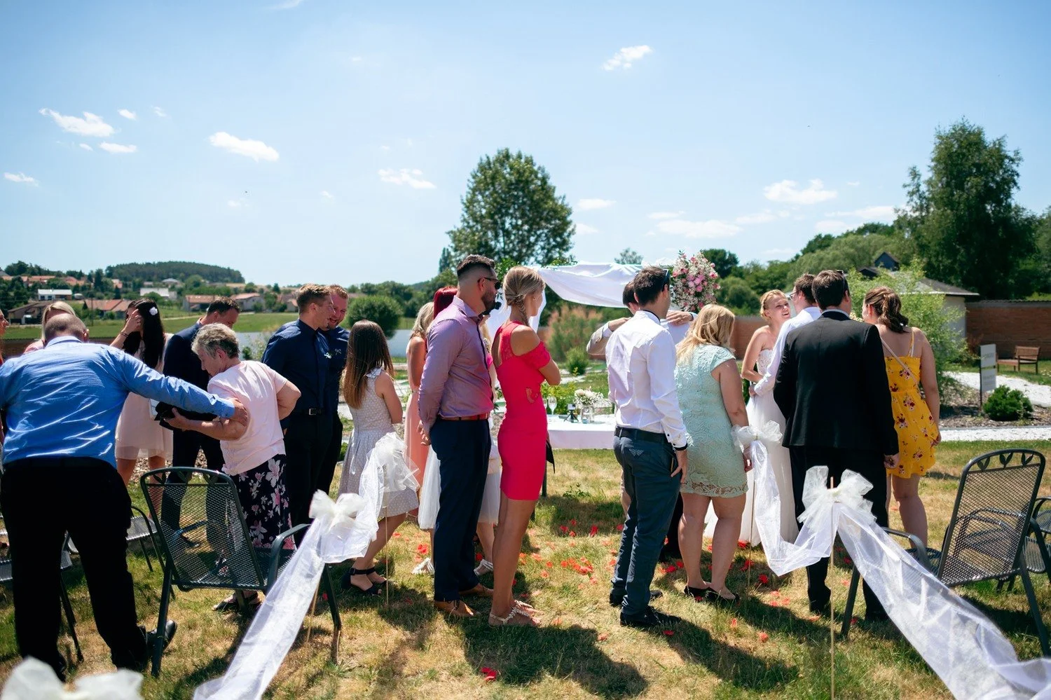 Hosté se za slunečného dne setkávají mezi židlemi a bílou výzdobou obřadní uličky.