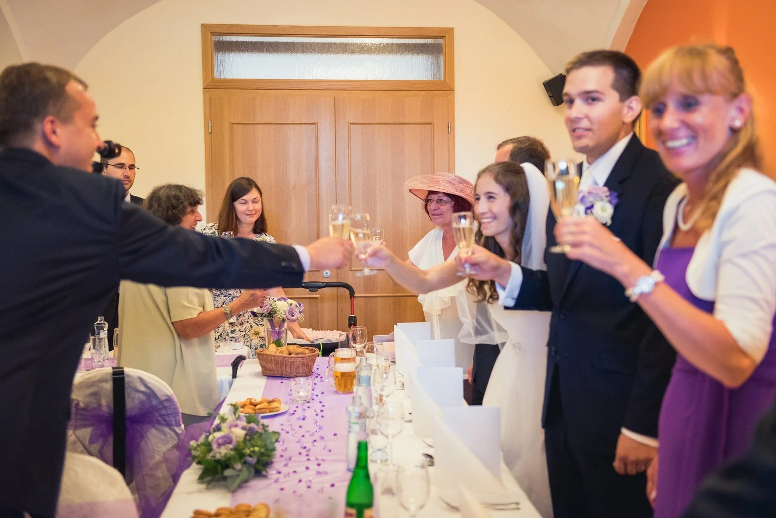 Několik lidí drží vysoko sklenky v živém společném přípitku přes šířku stolu.