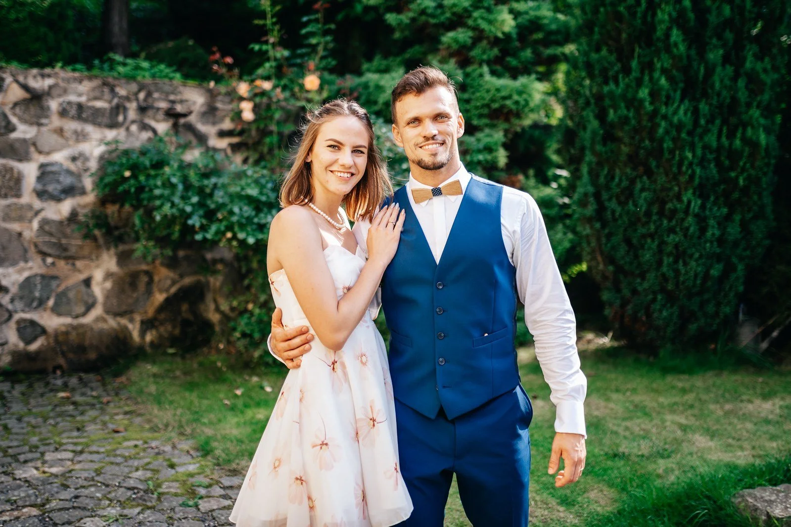 Dva mladí hosté pózují vedle sebe před kamennou zídkou.