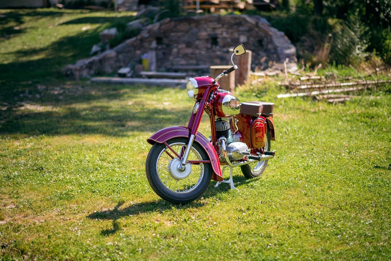 Červený historický motocykl stojí na slunci před kamennou zídkou.