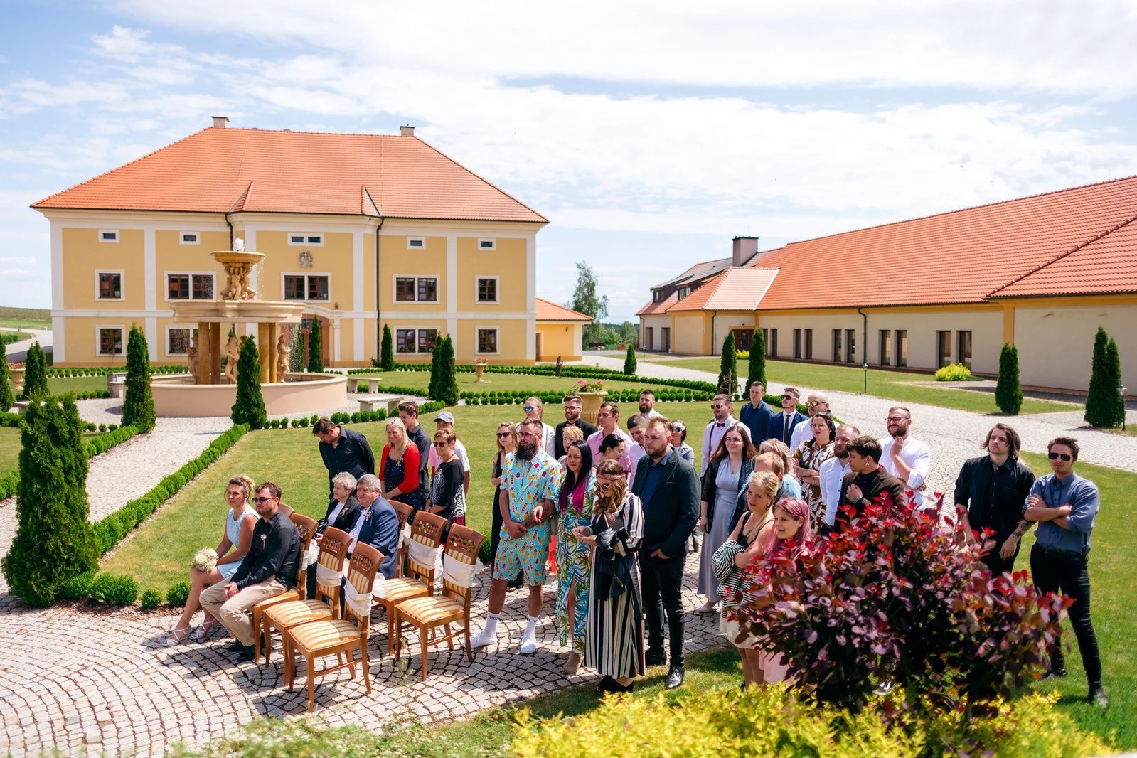 Klára přichází k venkovnímu obřadu v doprovodu muže v tmavém obleku.