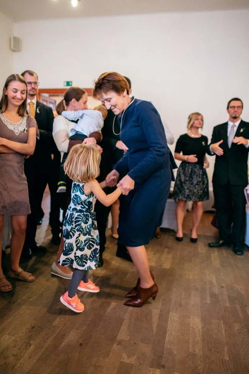 Muž v modrém obleku se sklání k malé dívce uprostřed parketu, zatímco kolem stojí hosté.