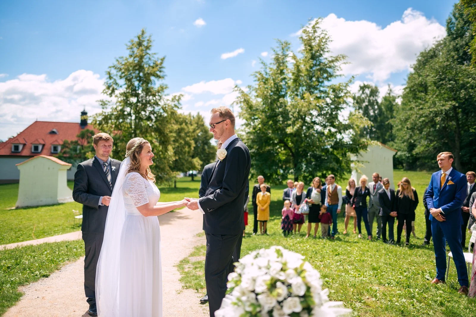 Žena v bílých šatech kráčí po cestě zavěšená do muže v tmavém obleku.