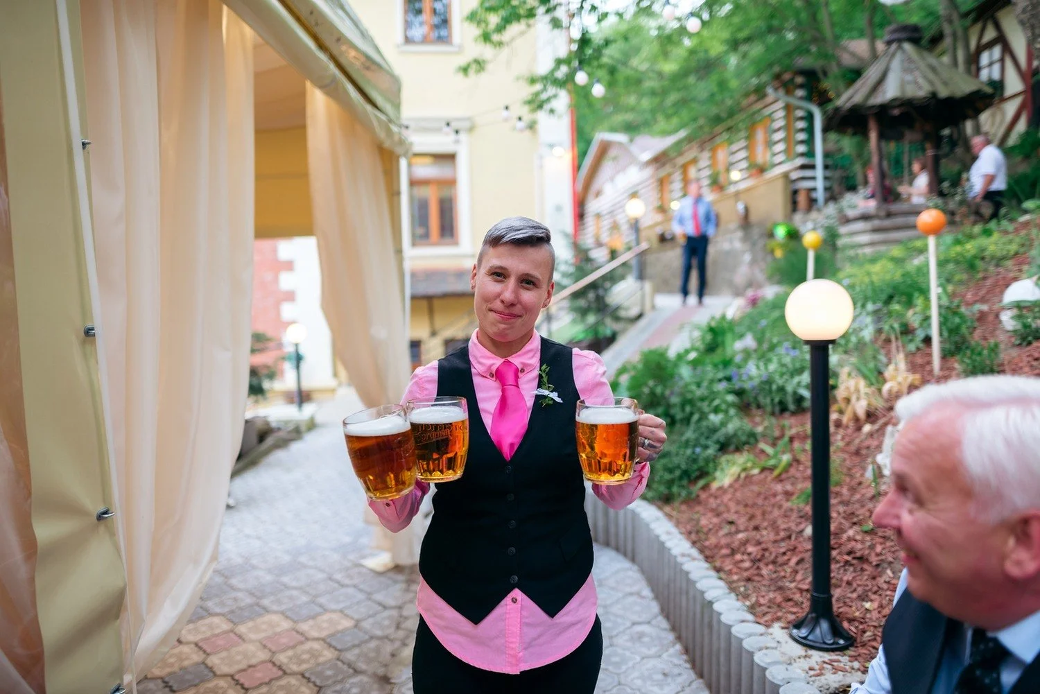 Číšník v růžové vestě přináší dvě sklenice piva zahradní uličkou mezi svatebními hosty.