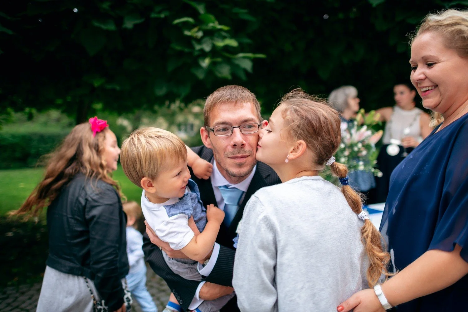 Muž s brýlemi sedí mezi dětmi během venkovního setkání hostů.