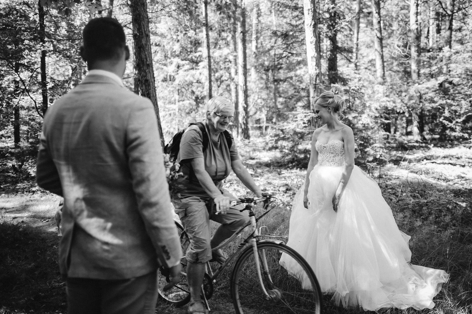 Černobílá momentka cyklisty projíždějícího mezi slavnostně oblečenou dvojicí v lese.