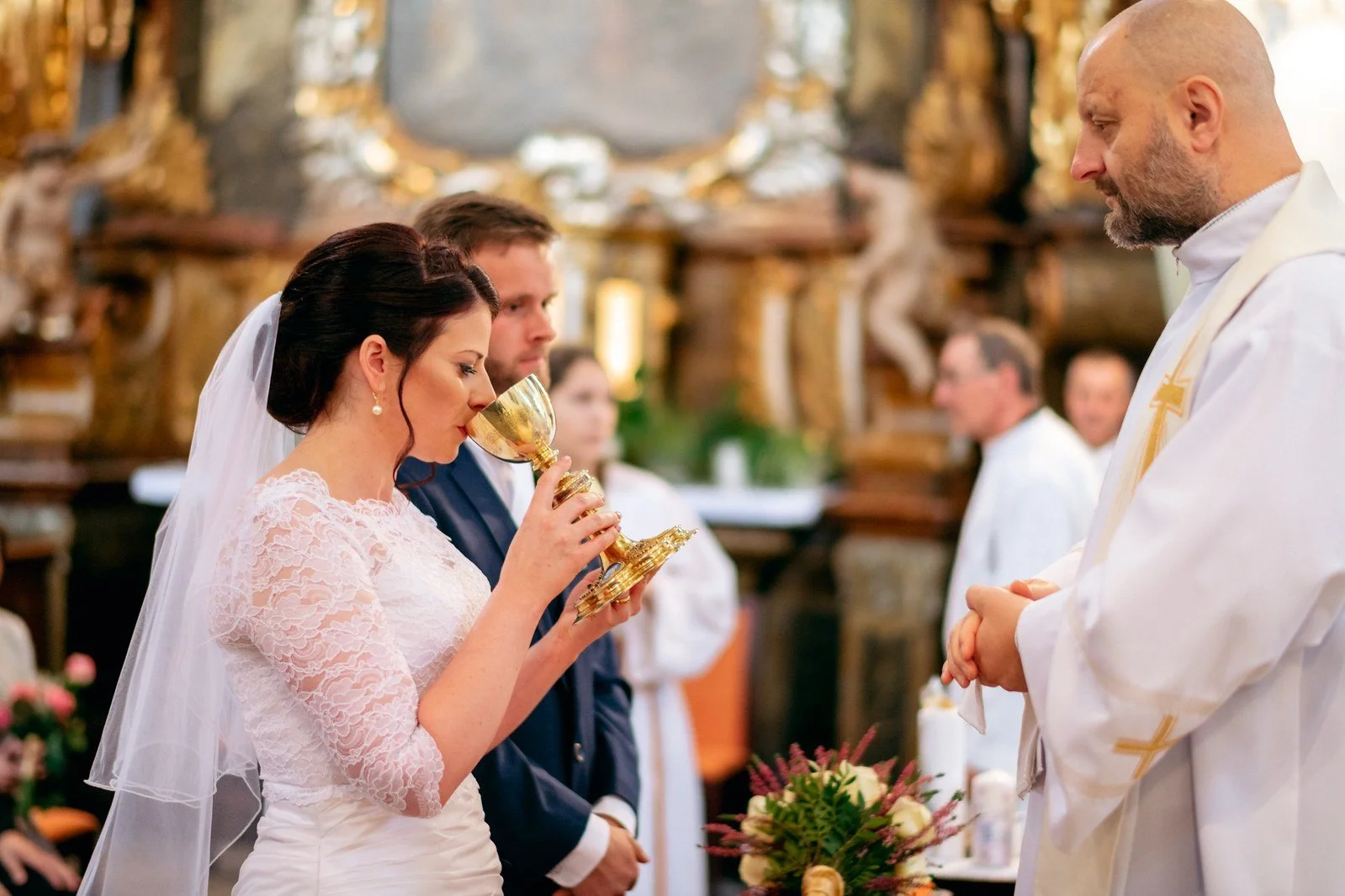 Žena se závojem pije ze zlatého kalicha podávaného mužem ve světlém rouchu.
