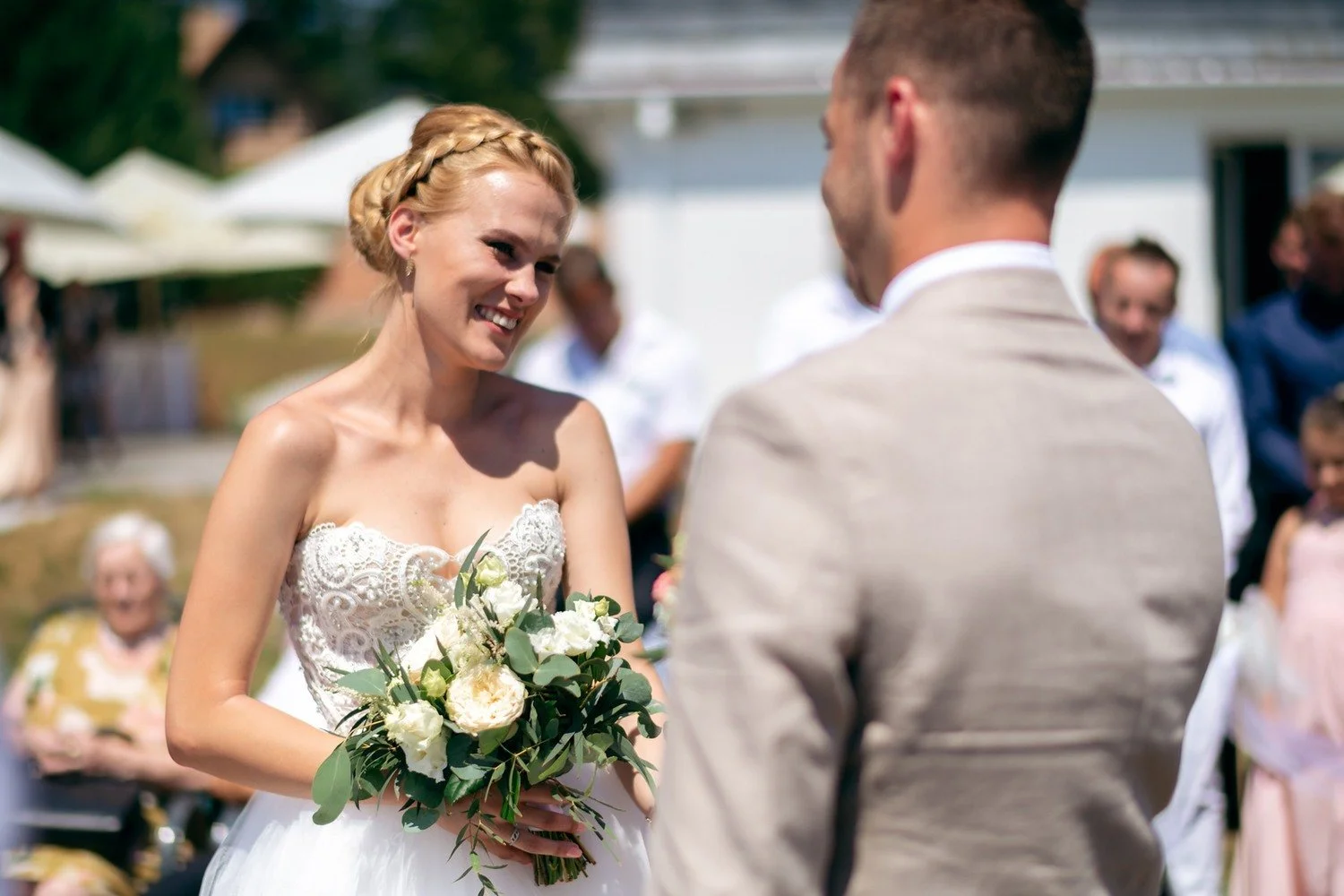 Lucie s kyticí hledí na ženicha ve světlém obleku během venkovního obřadu.