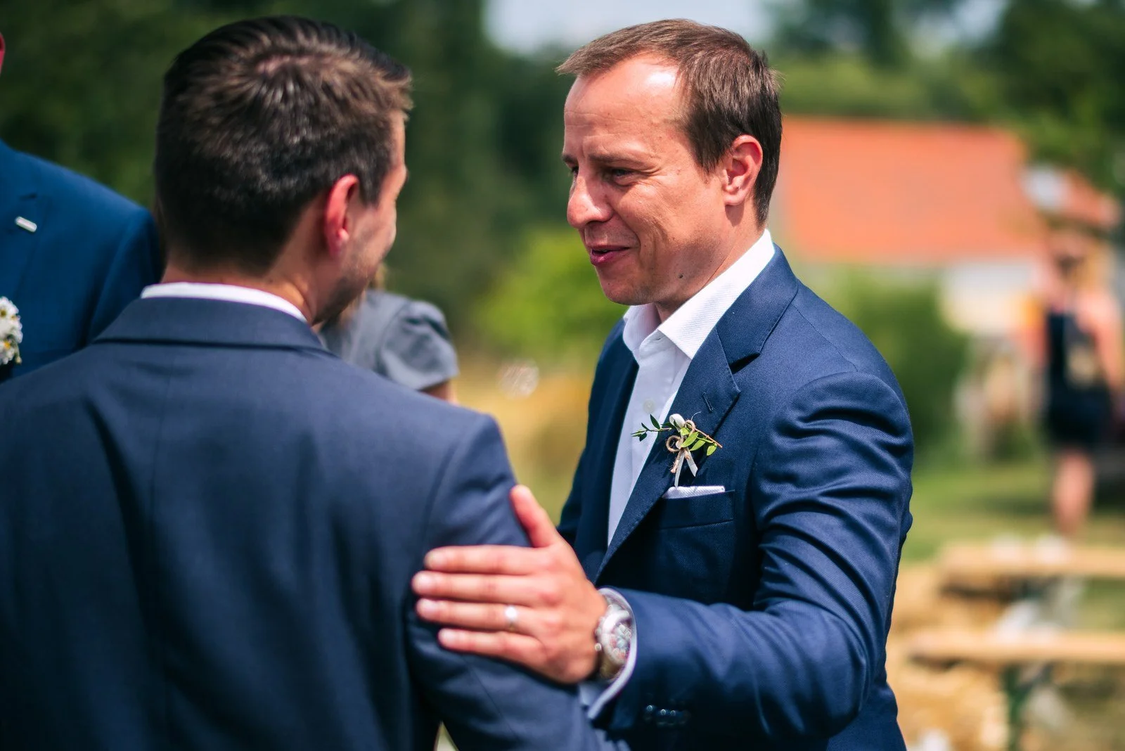 Dva muži v oblecích se zdraví zblízka a jeden pokládá druhému ruku na rameno.