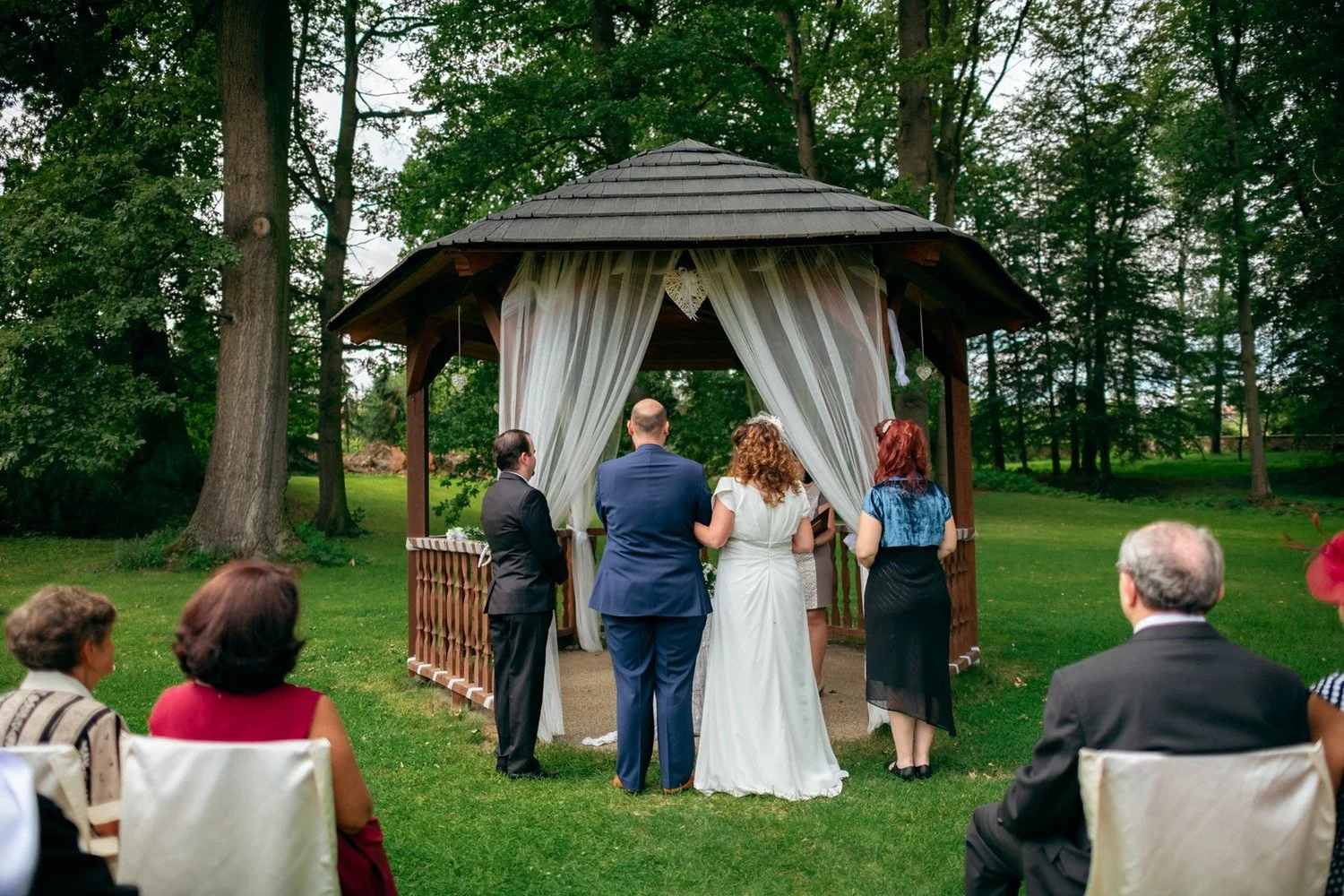 Pohled zezadu zachycuje pár a hosty během obřadu v altánu.