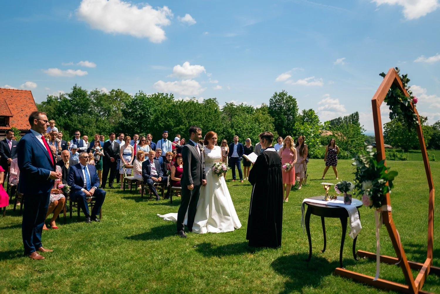 Široký pohled na nevěstu a ženicha před oddávající, hosty a květinami zdobeným dřevěným obloukem.