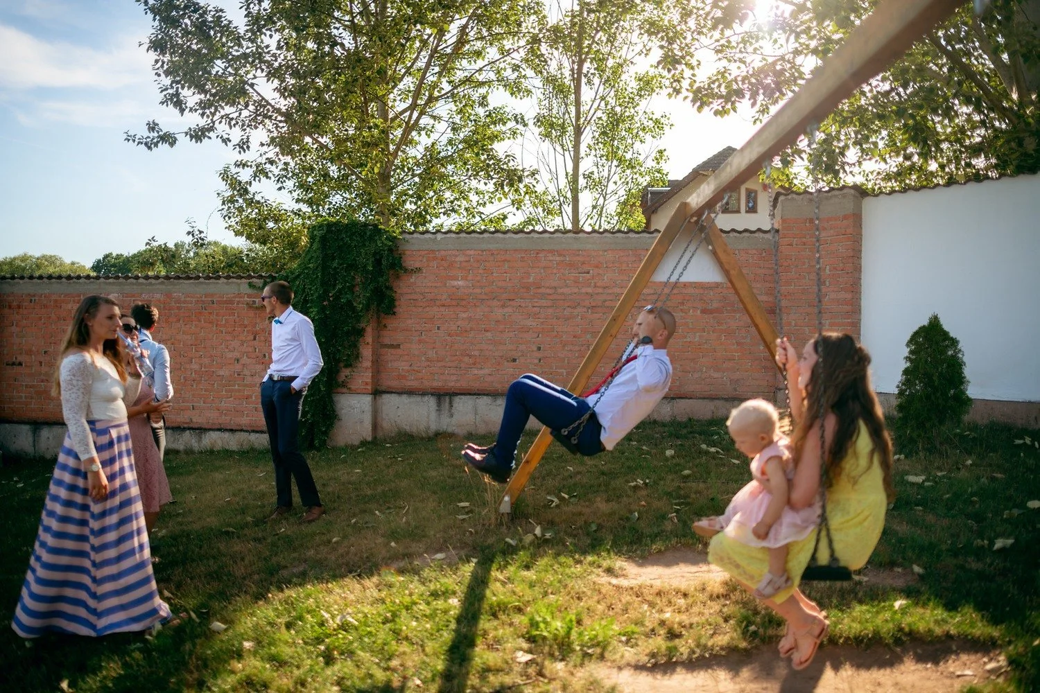 Dítě ve žlutém stojí na trávníku u modré houpačky, zatímco opodál přihlížejí hosté.