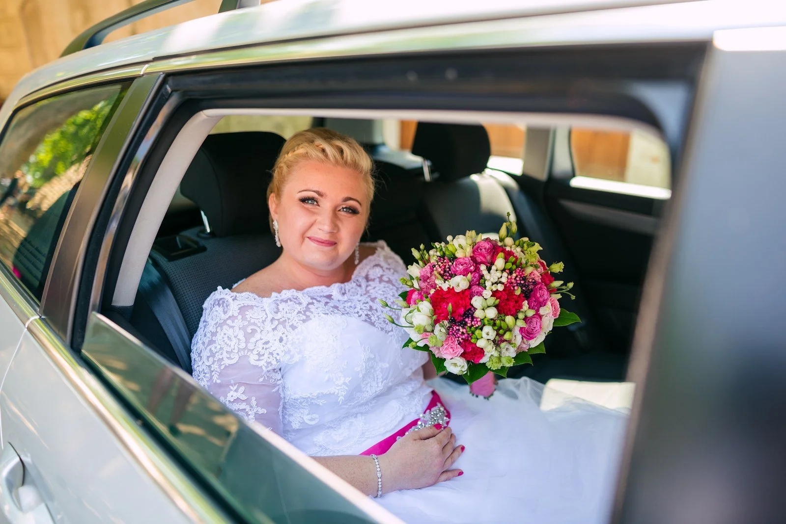 Žena v bílých šatech drží barevnou kytici u otevřeného okna auta.