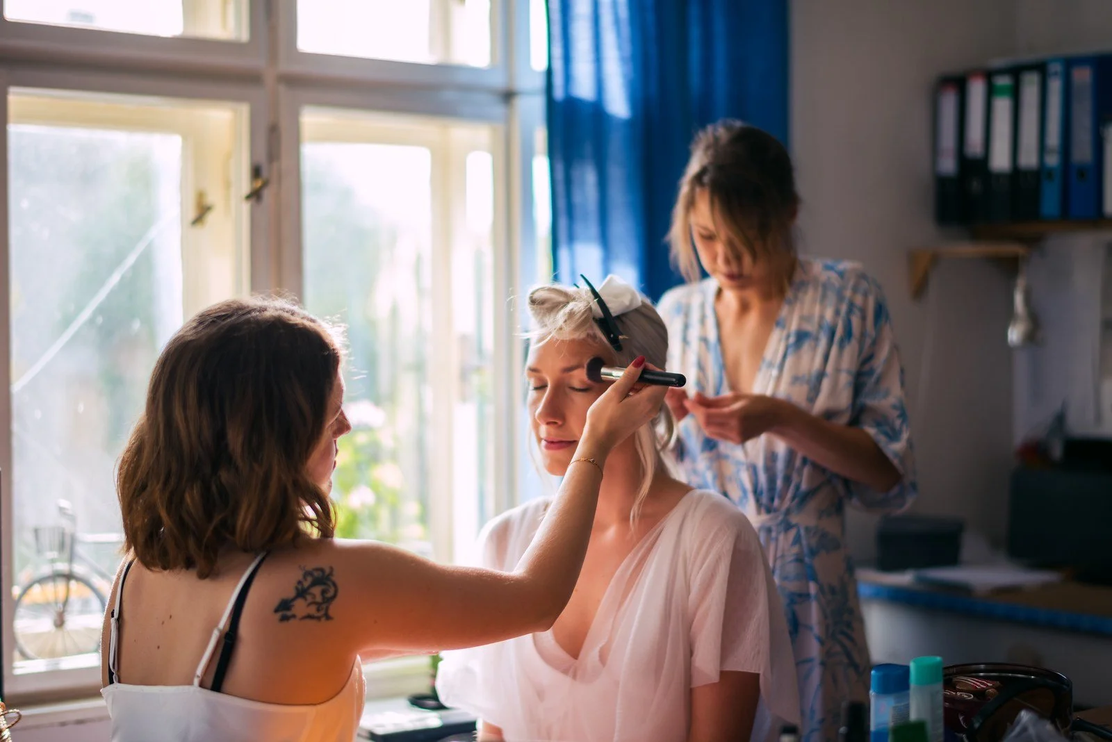 Dvě ženy upravují vlasy a líčení blond ženě v růžovém županu u světlého okna.