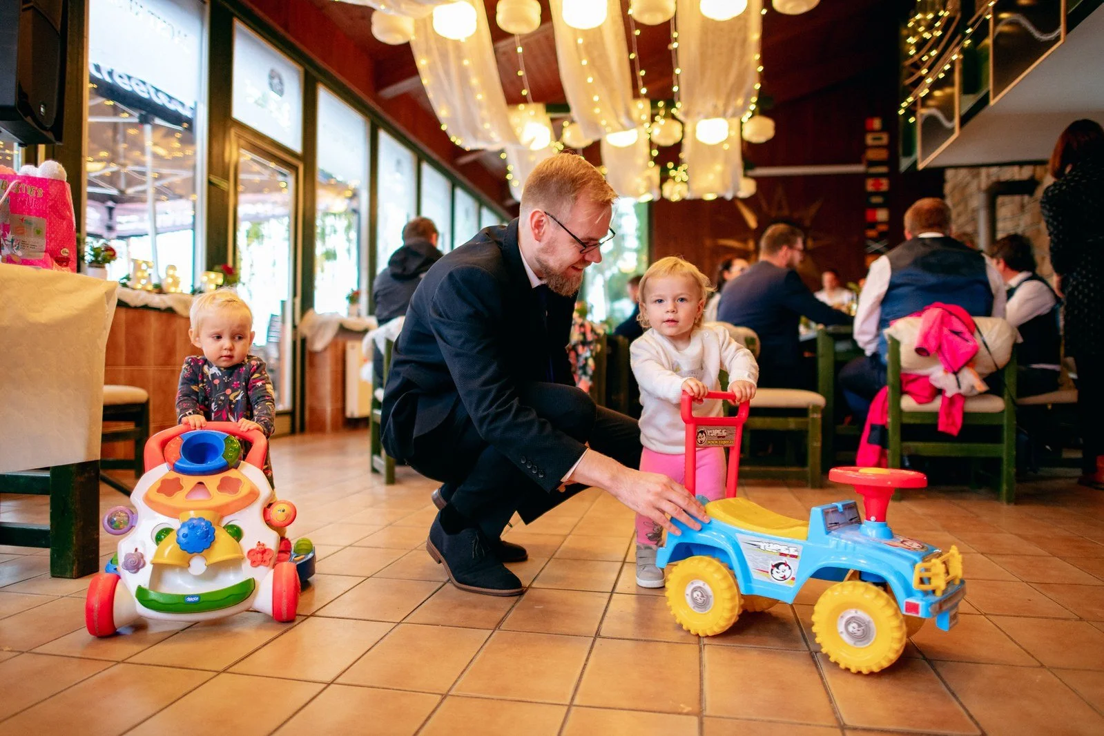 Dvě malé děti sedí na barevných odrážedlech mezi hosty v osvětleném sále.