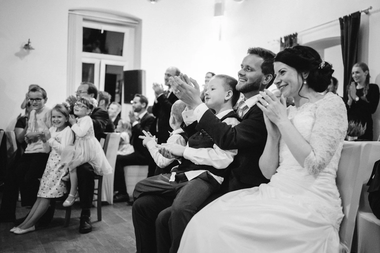 Černobílá fotografie zachycuje sedící dvojici s dítětem na klíně při společném potlesku.