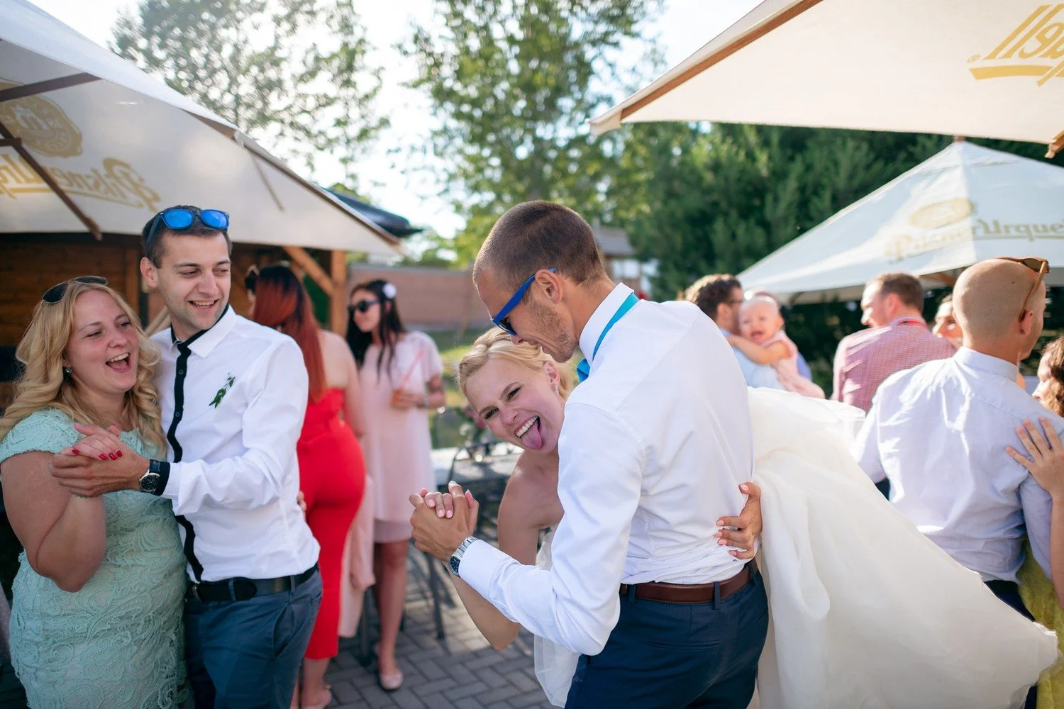 Rozesmátá skupina hostů tančí na terase, muž v bílé košili se sklání s nápojem v ruce.