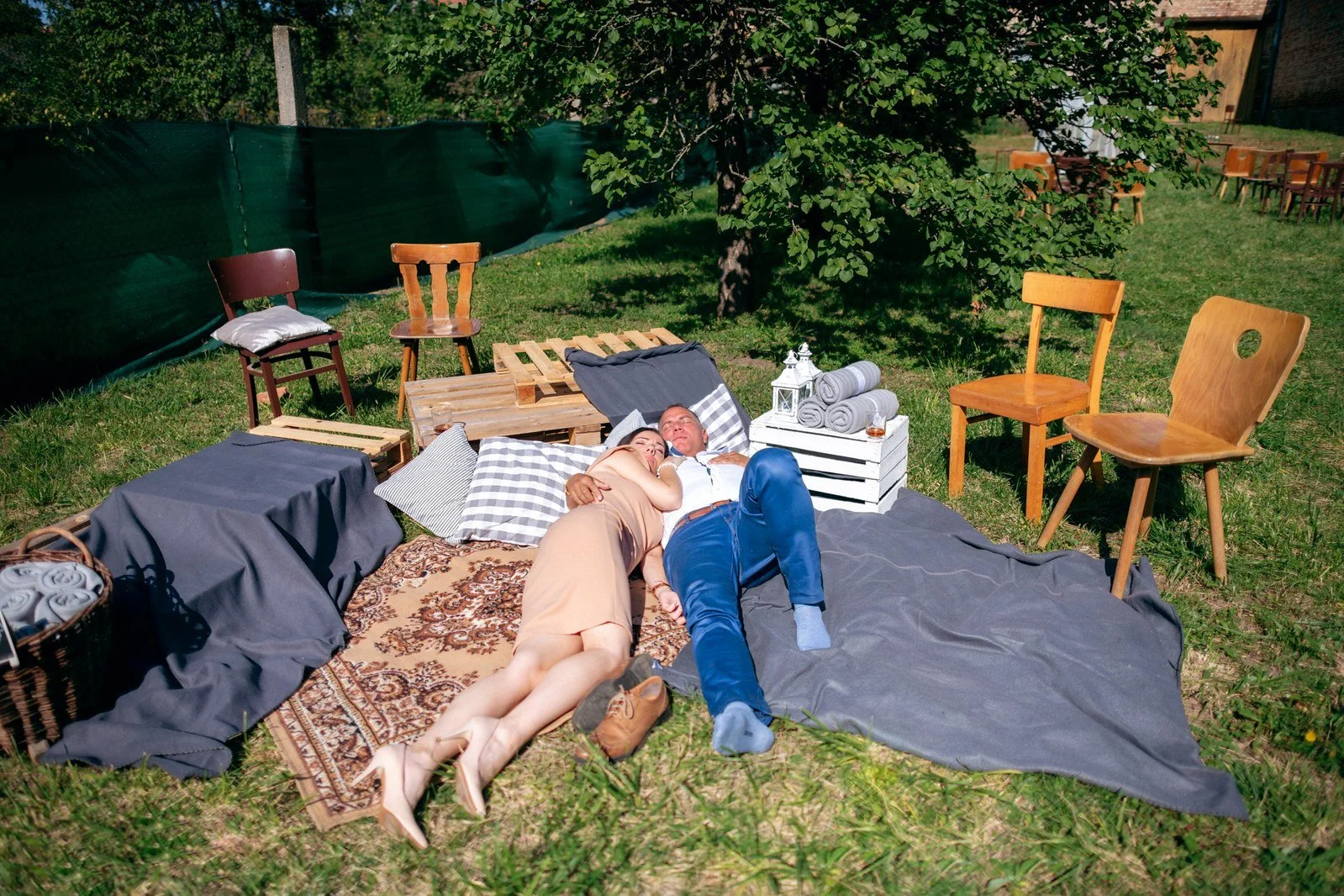Dvě osoby leží vedle sebe na dekách a polštářích v zahradním odpočinkovém koutku obklopeném židlemi.