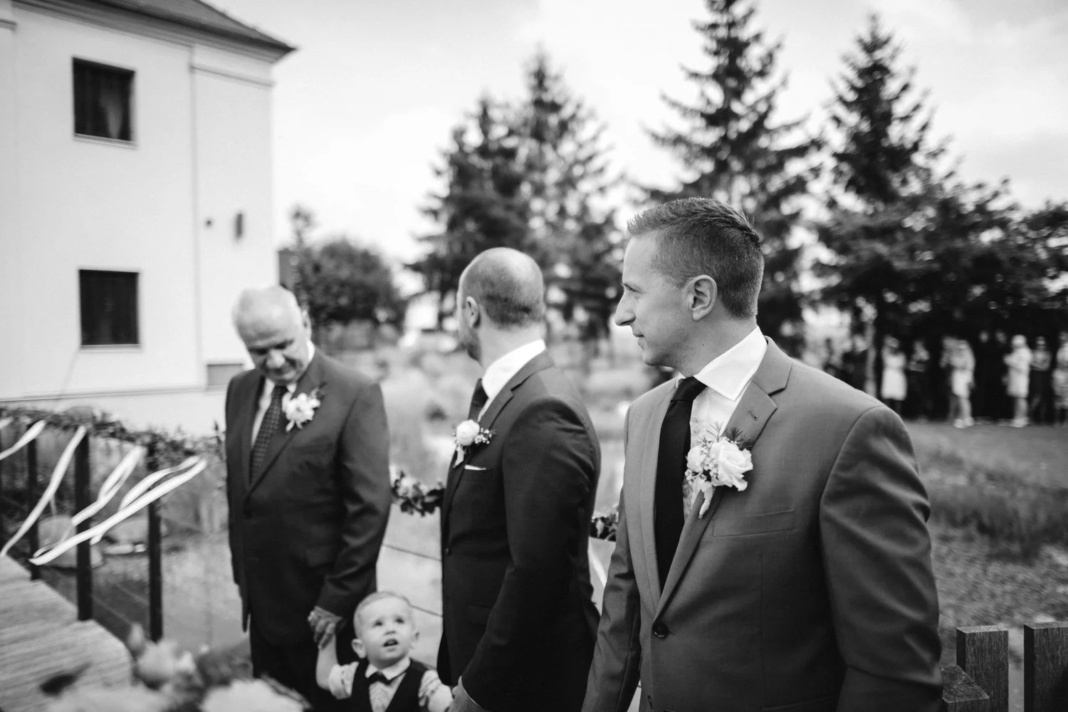 Černobílý moment ženicha s hosty a malým chlapcem před začátkem obřadu.