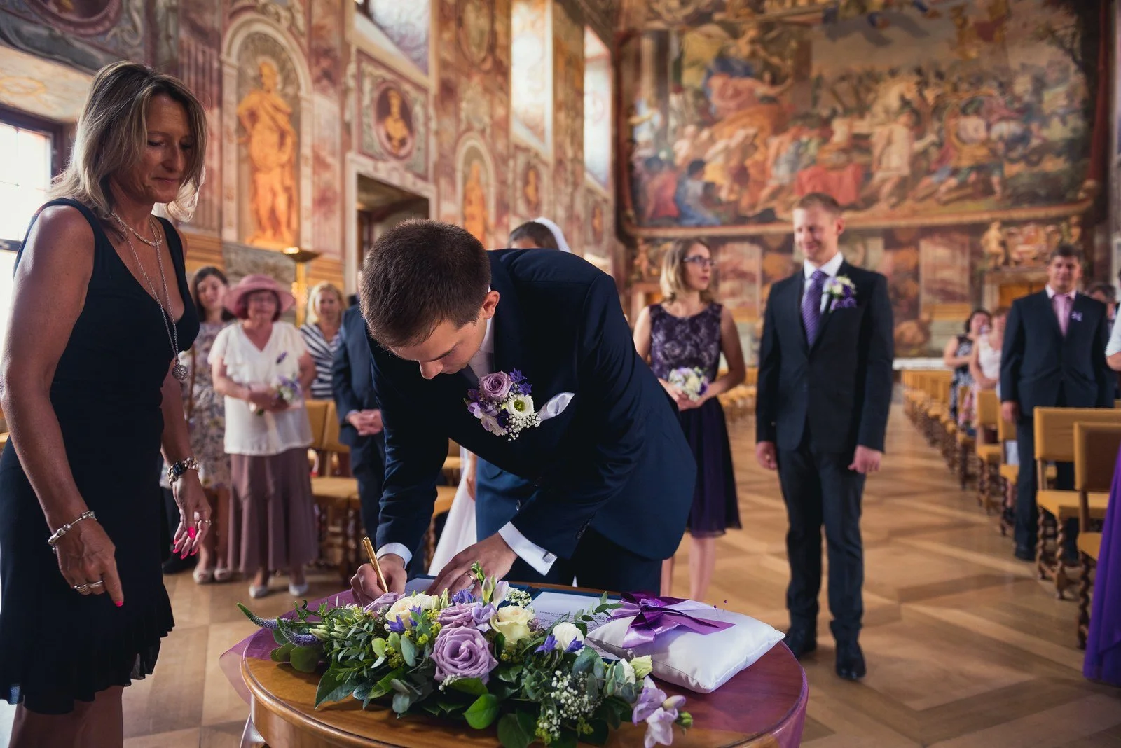 Žena v bílých šatech se sklání k podpisu u stolku s květinami, zatímco několik osob přihlíží.