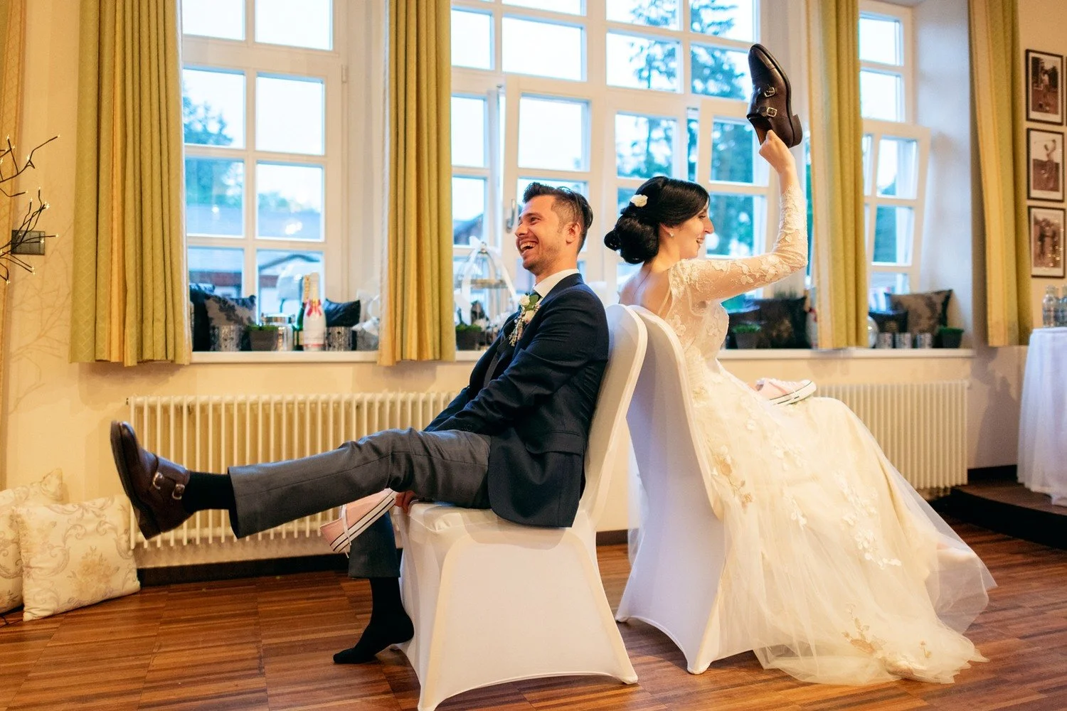 Nevěsta a ženich sedí zády k sobě na židlích a každý zvedá nad hlavu jednu pánskou botu.