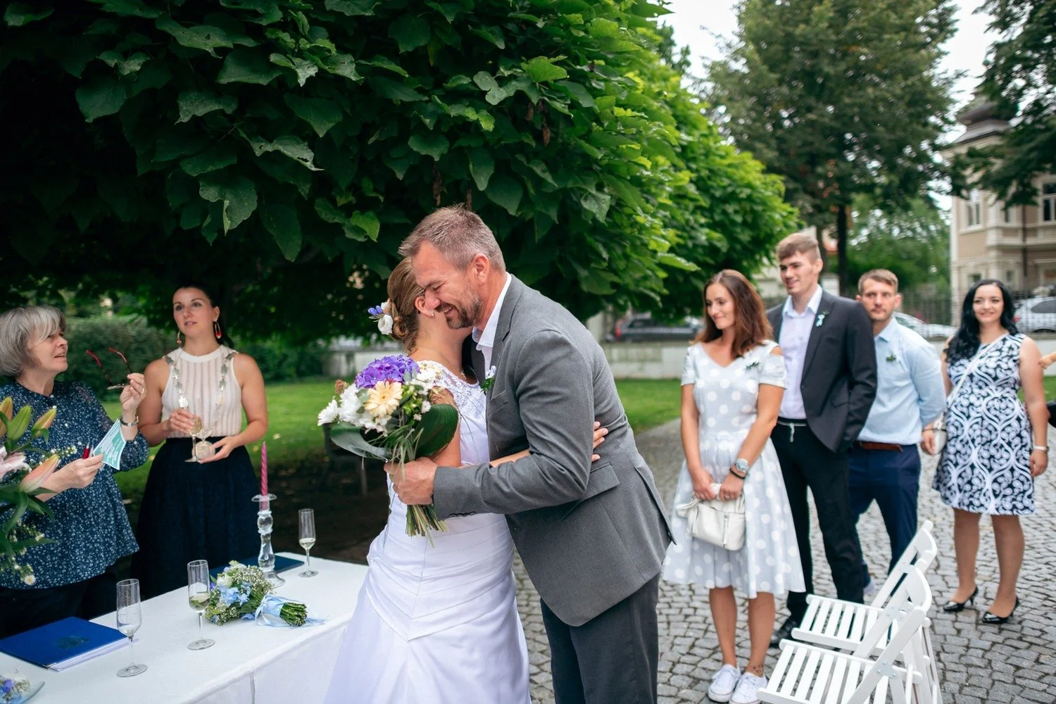 Nevěsta s kyticí objímá muže v šedém obleku.
