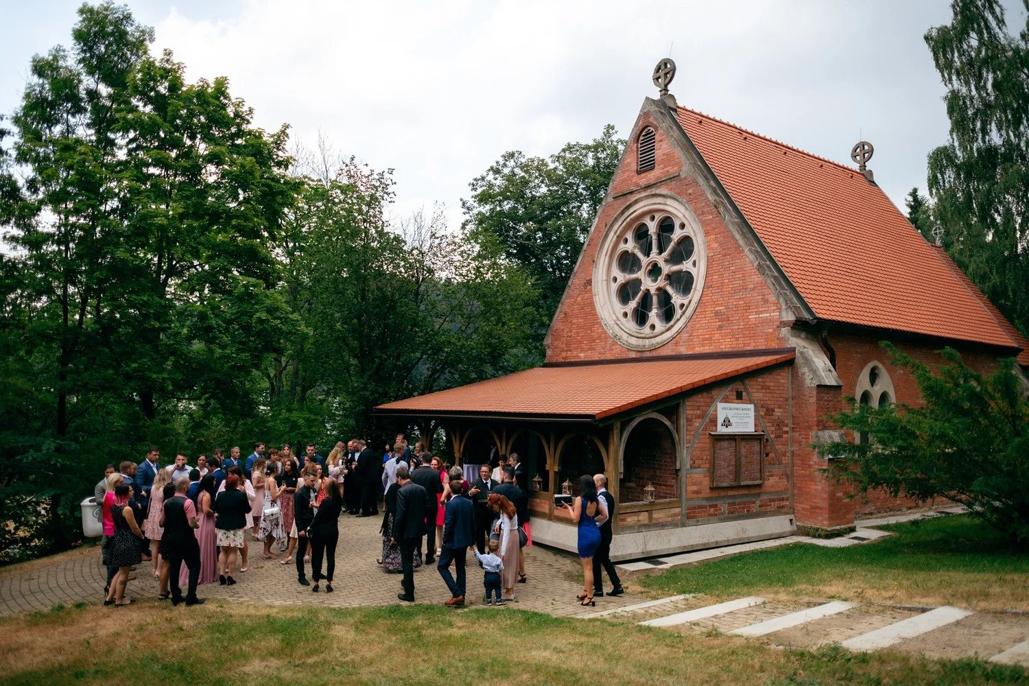 Skupina slavnostně oblečených lidí se shromáždila na prostranství před cihlovým kostelem.