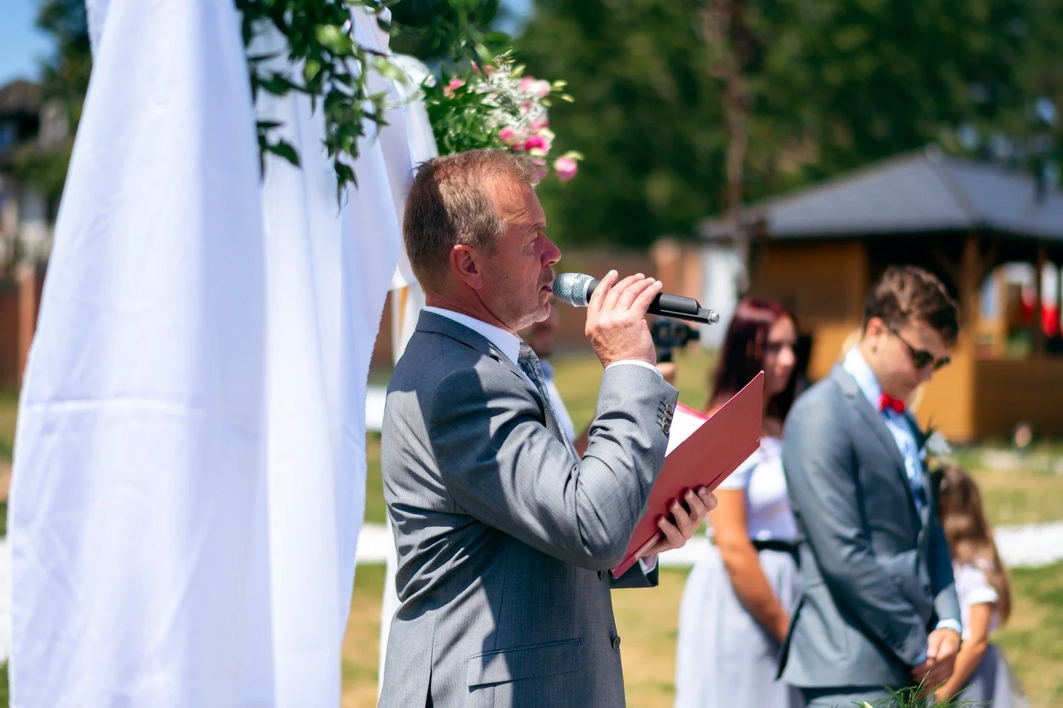 Muž v šedém obleku mluví do mikrofonu před hosty u obřadní slavobrány.