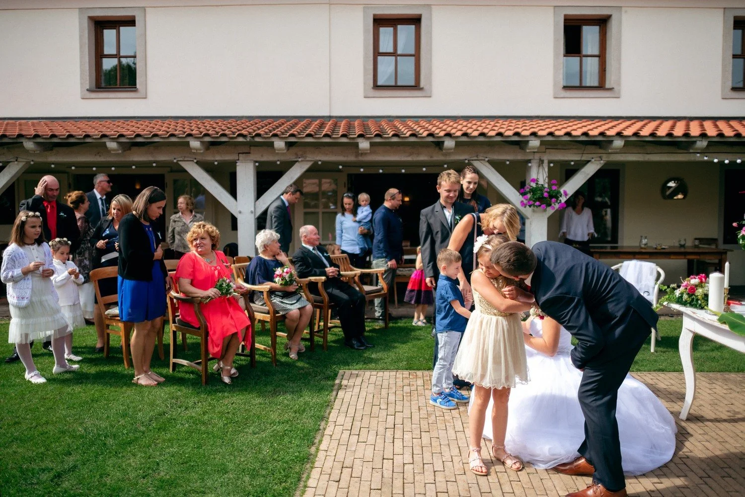 Muž v obleku se sklání k malé dívce stojící na trávníku před přihlížejícími lidmi.