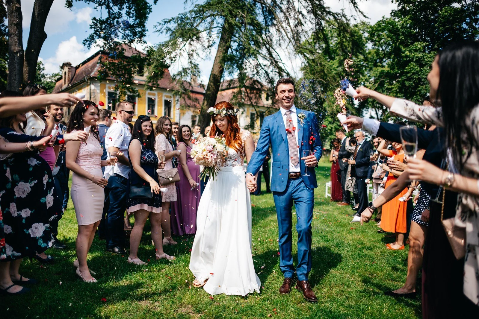 Novomanželé procházejí uličkou mezi hosty s rukama zvednutýma nad hlavou.