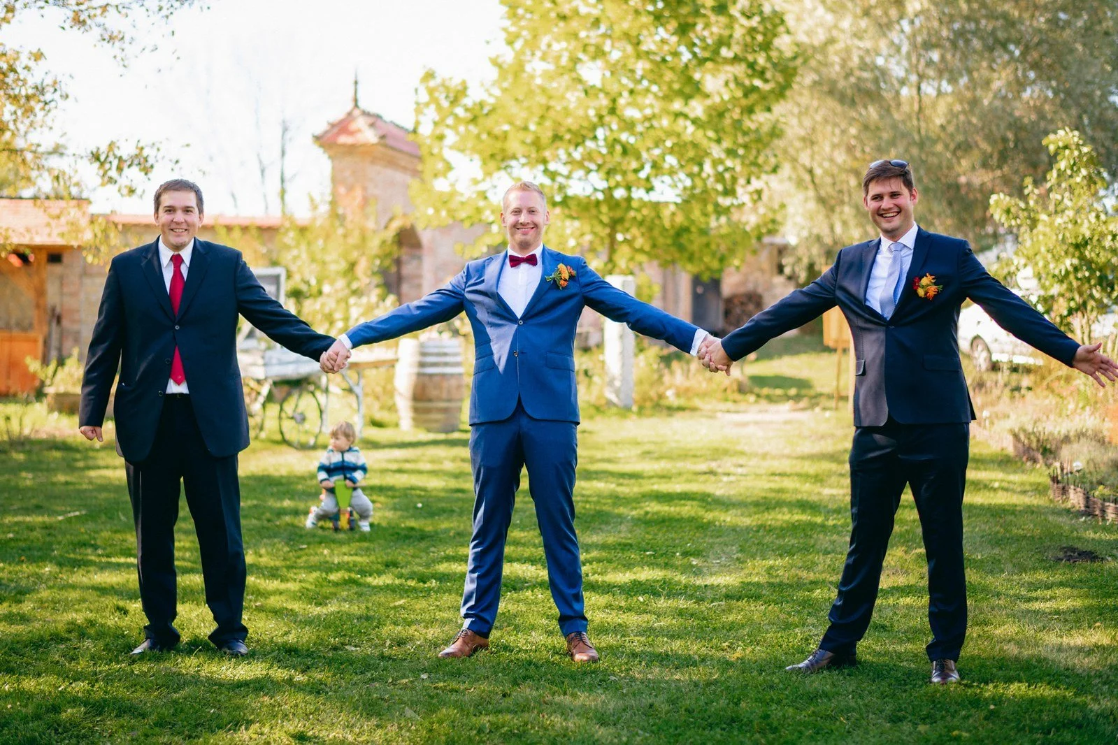 Tři muži v rozestupu a s rozpaženými pažemi narušují formálnost skupinových snímků humorem.