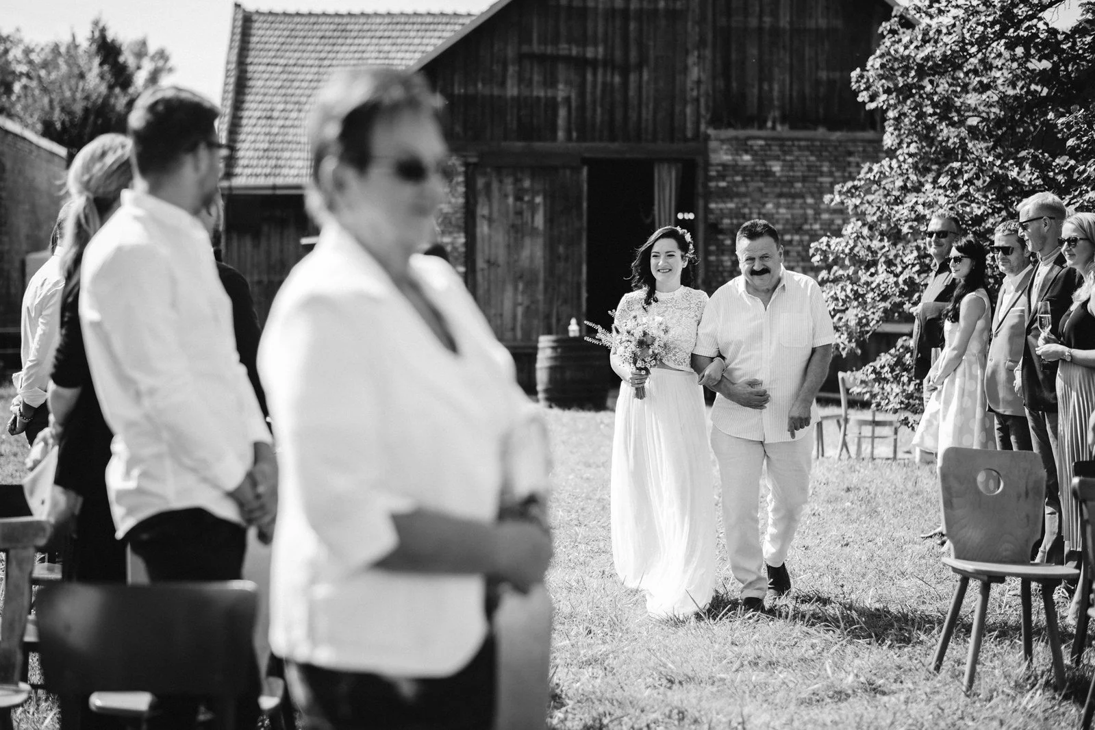 Černobílý záběr přes rozostřené stojící hosty ukazuje ženu v dlouhých bílých šatech přicházející s mužem.