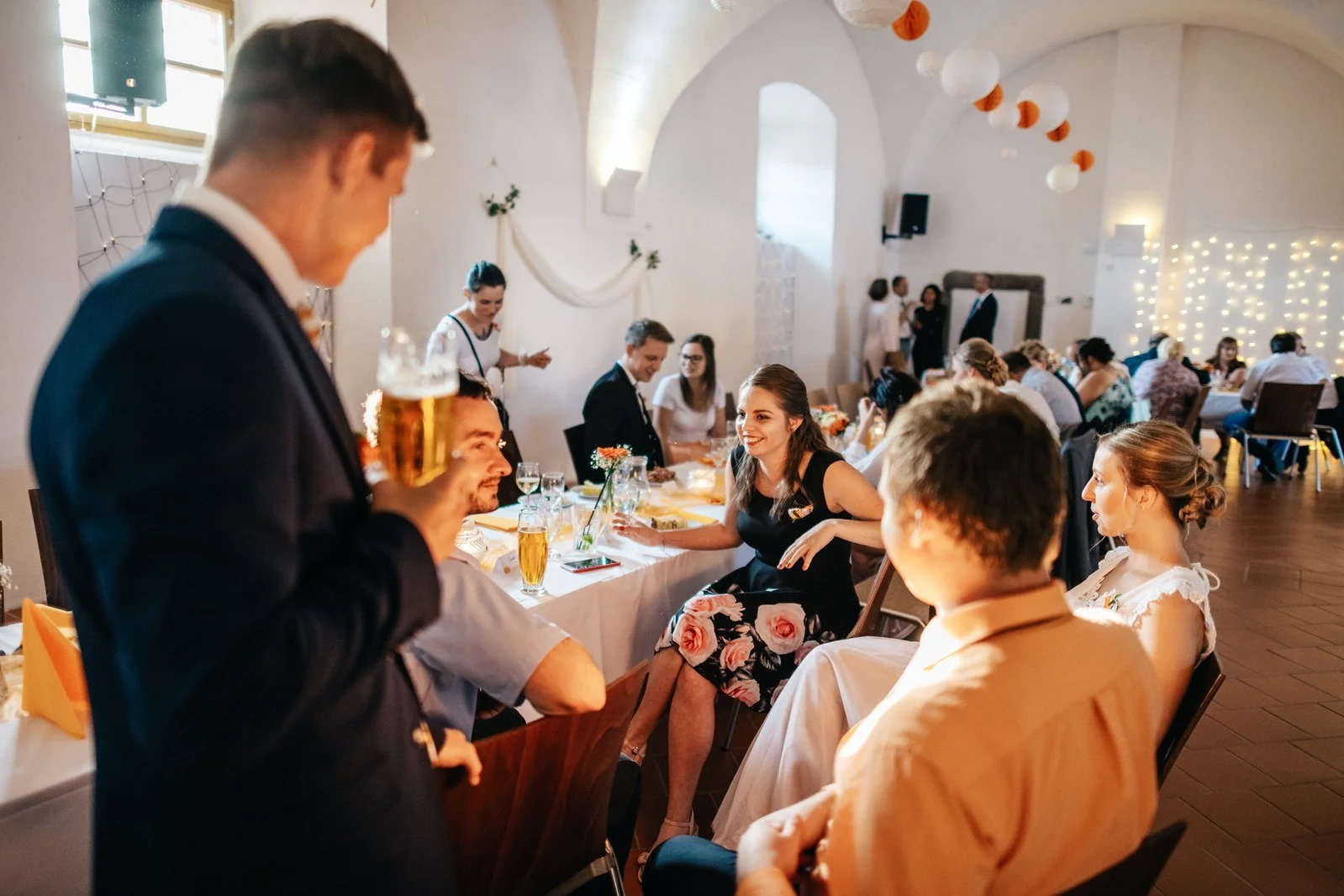Hosté sedí u stolů a otáčejí se k ženichovi stojícímu poblíž.