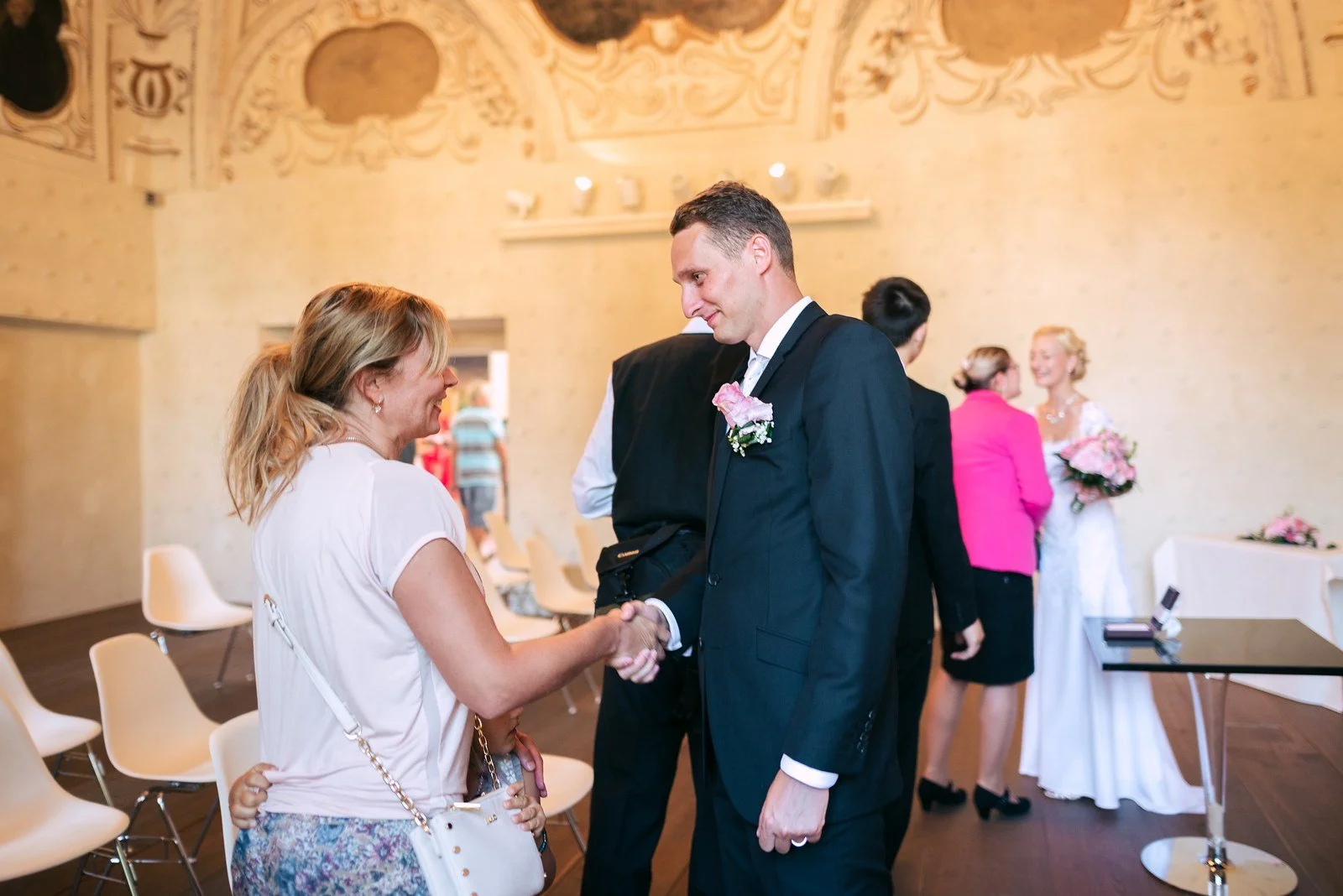 Dvojice stojí vedle sebe s hosty v pozadí obřadní síně.