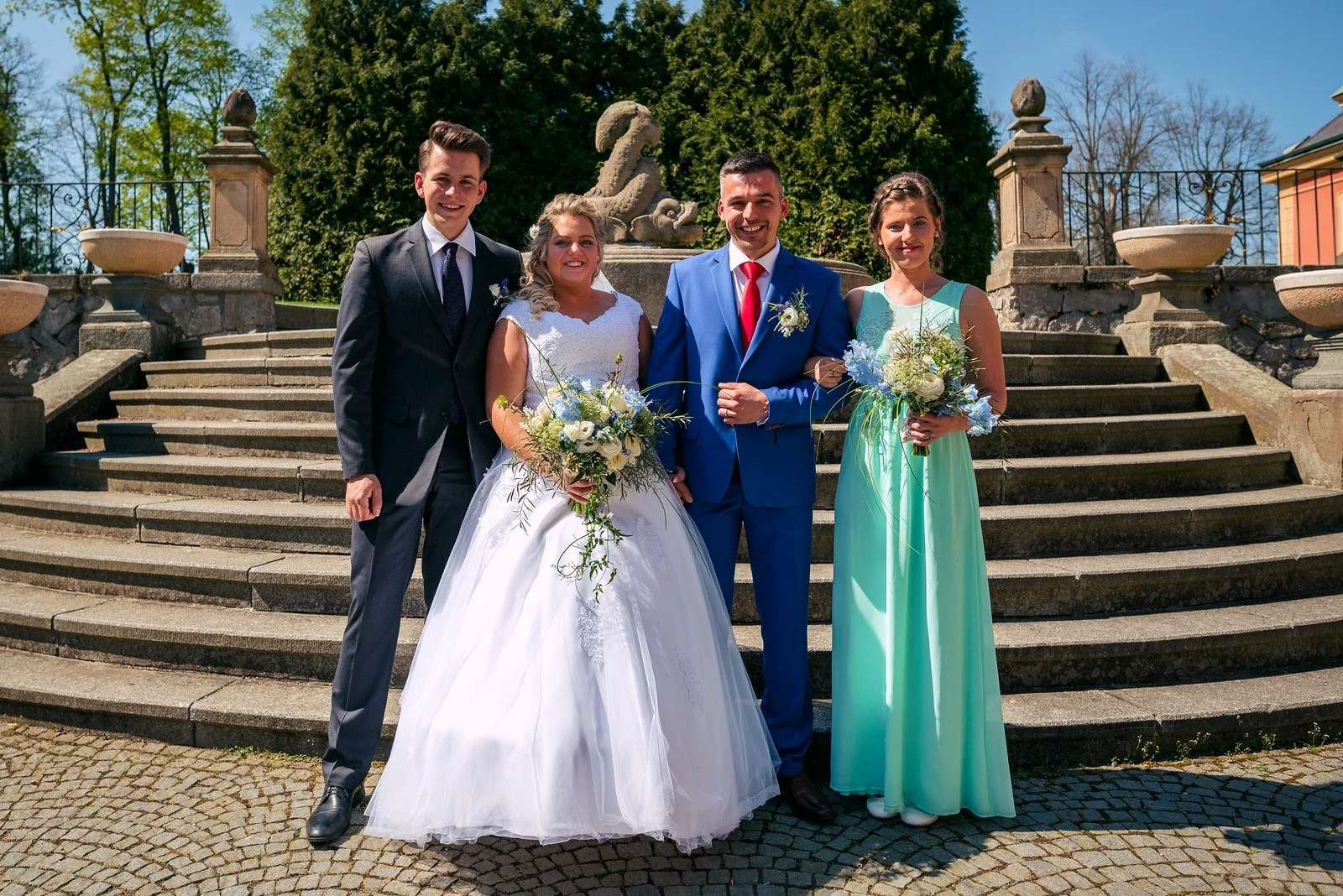 Barevně oblečená skupina hostů s novomanželi na schodech Zámku Dobříš.