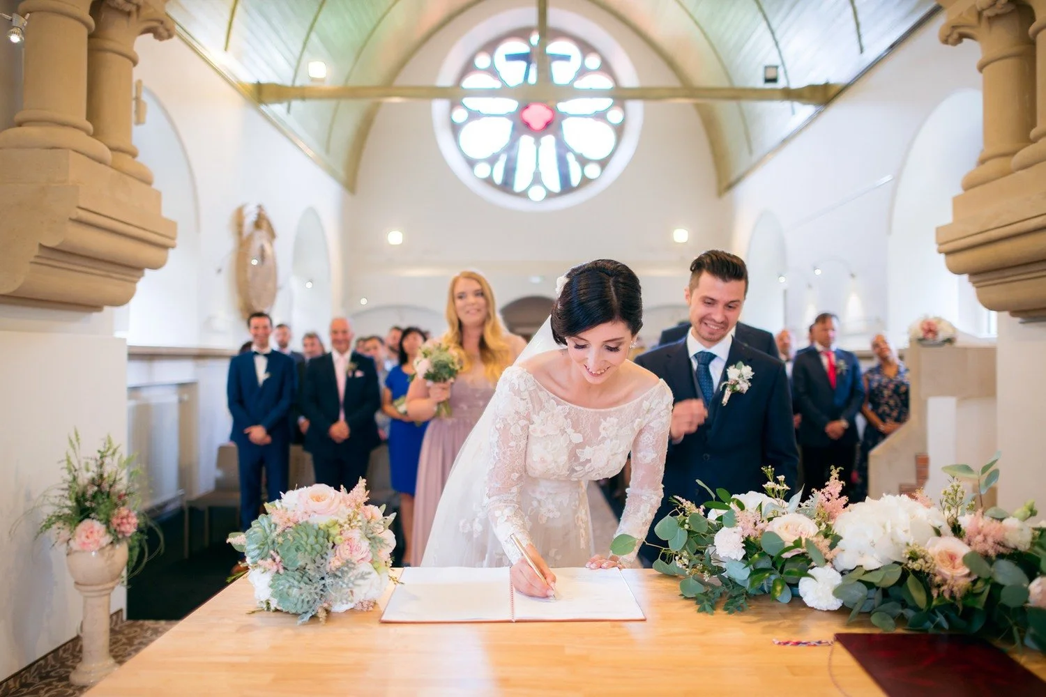Kateřina se sklání nad obřadním stolem a podepisuje otevřený dokument mezi květinovými aranžemi.