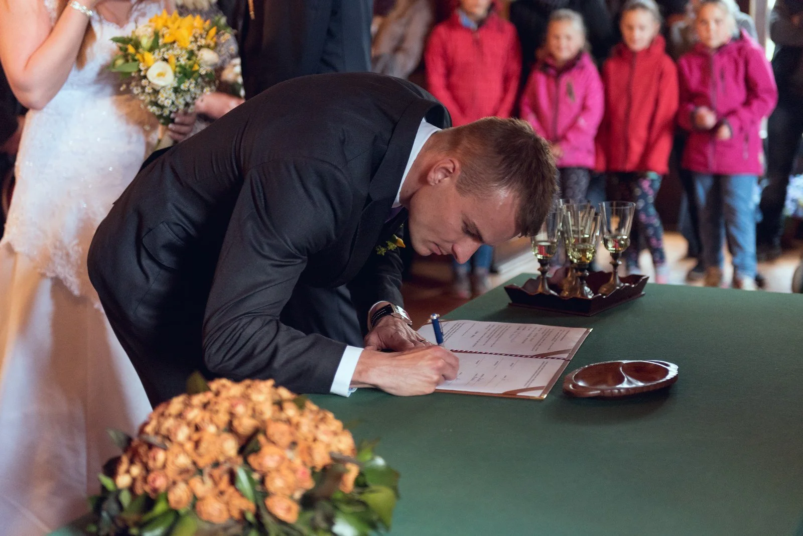 Ženich se sklání a podepisuje dokument na stole před řadou hostů.