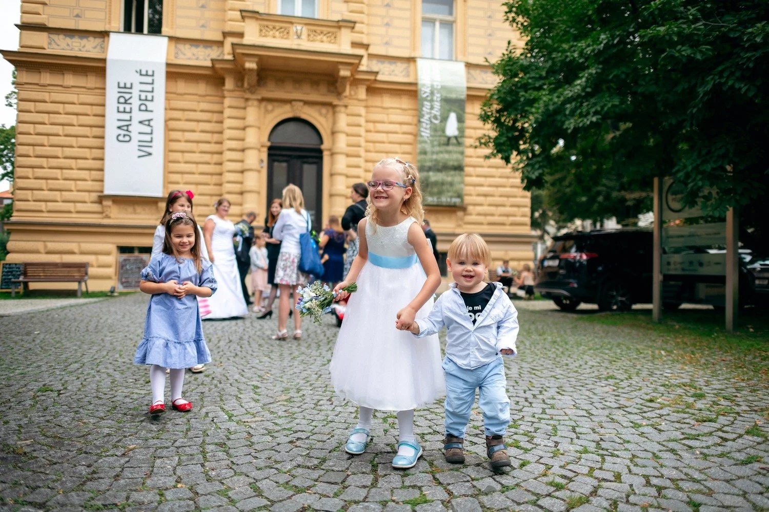 Žena v bílých šatech vede malého chlapce po dlážděné cestě před vilou.