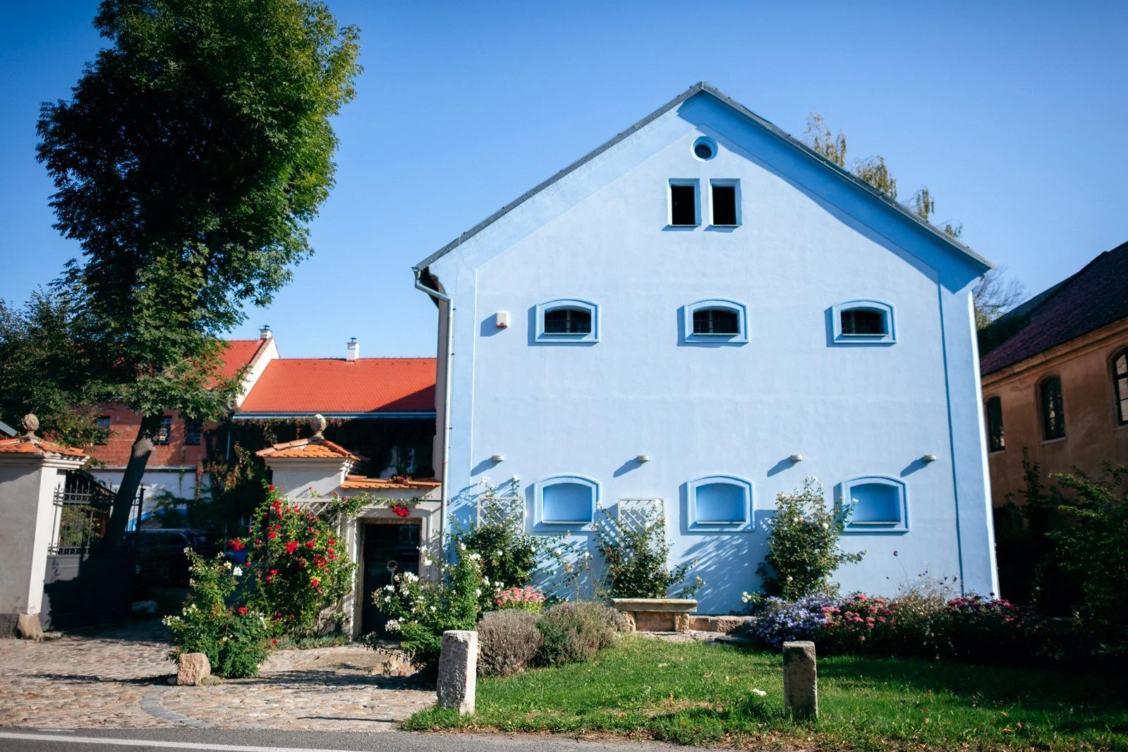 Světlý patrový dům s červenou střechou stojí za zelenou předzahrádkou pod modrou oblohou.