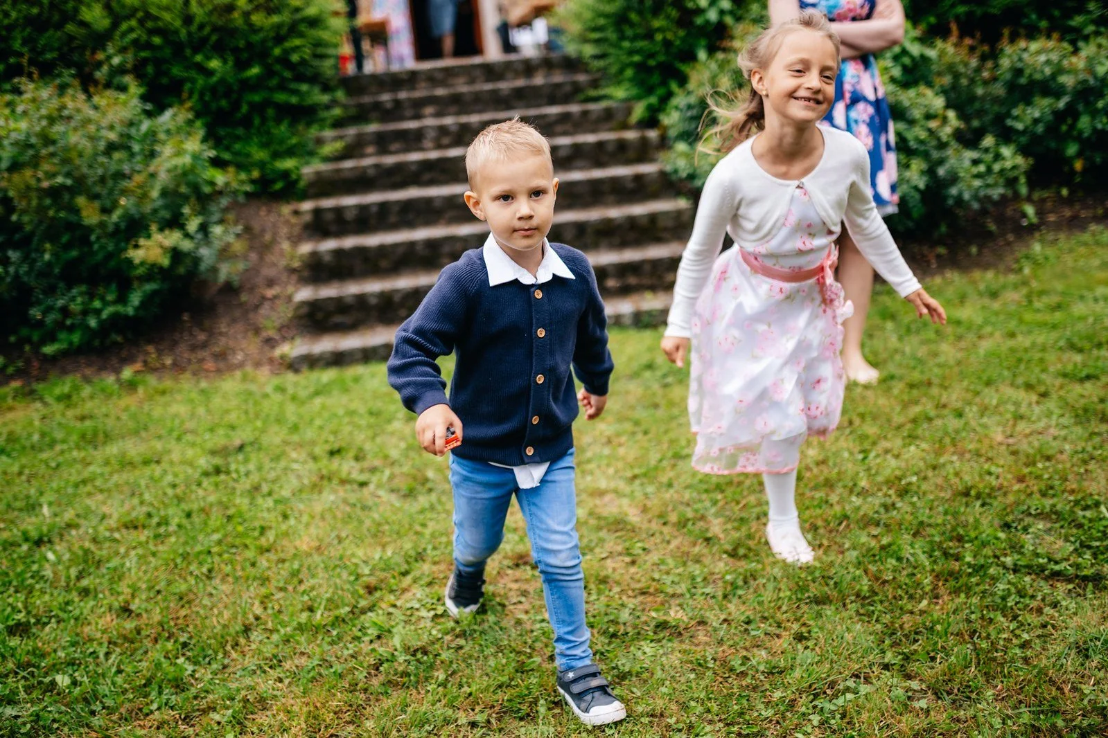 Chlapec v modrém saku a dívka ve světlých šatech běží vedle sebe po trávě v zahradě.